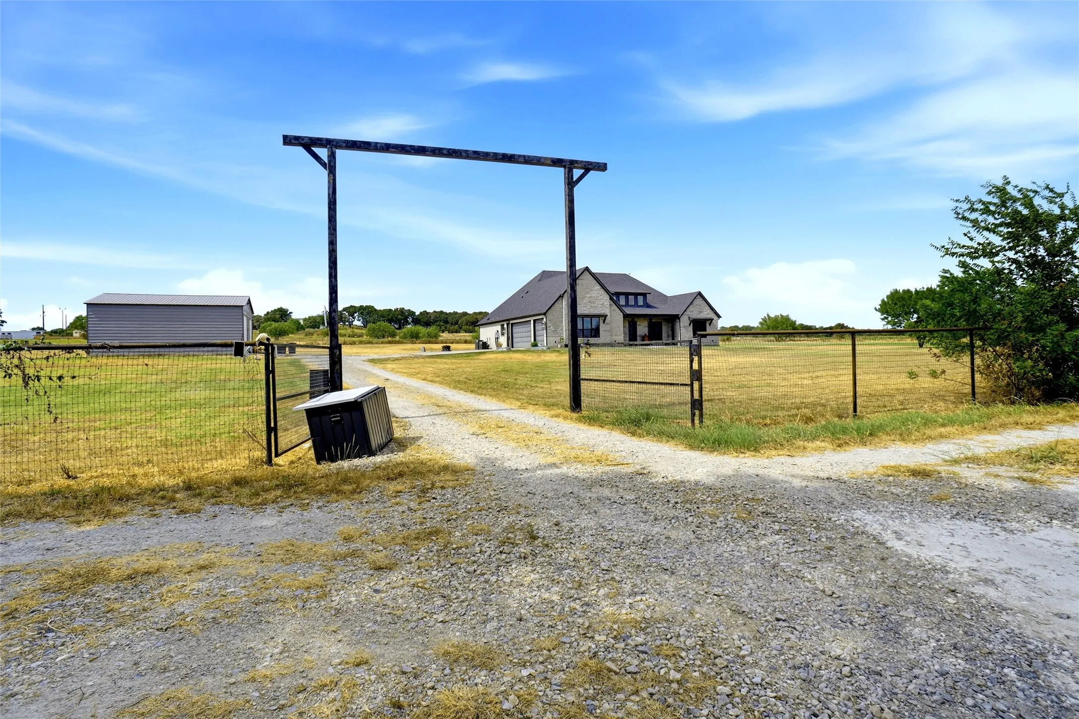 Gravel driveway leading to a stone exterior residence with a gray metal roof, featuring a detached metal-sided building and a fenced perimeter