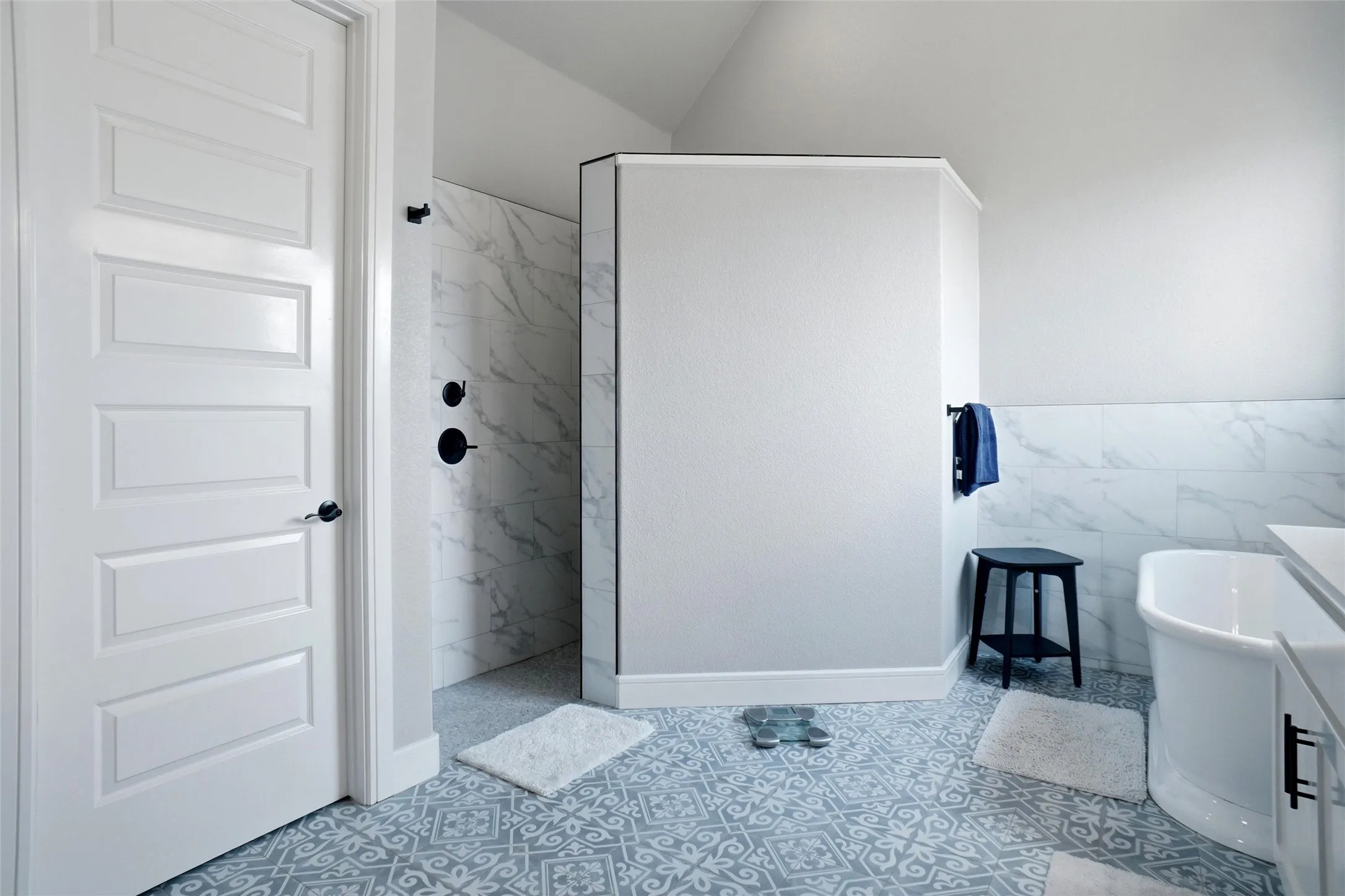 Patterned floor tiles, freestanding bathtub, walk-in shower with marble-style tile surround, matte black shower fixtures, and a paneled interior door