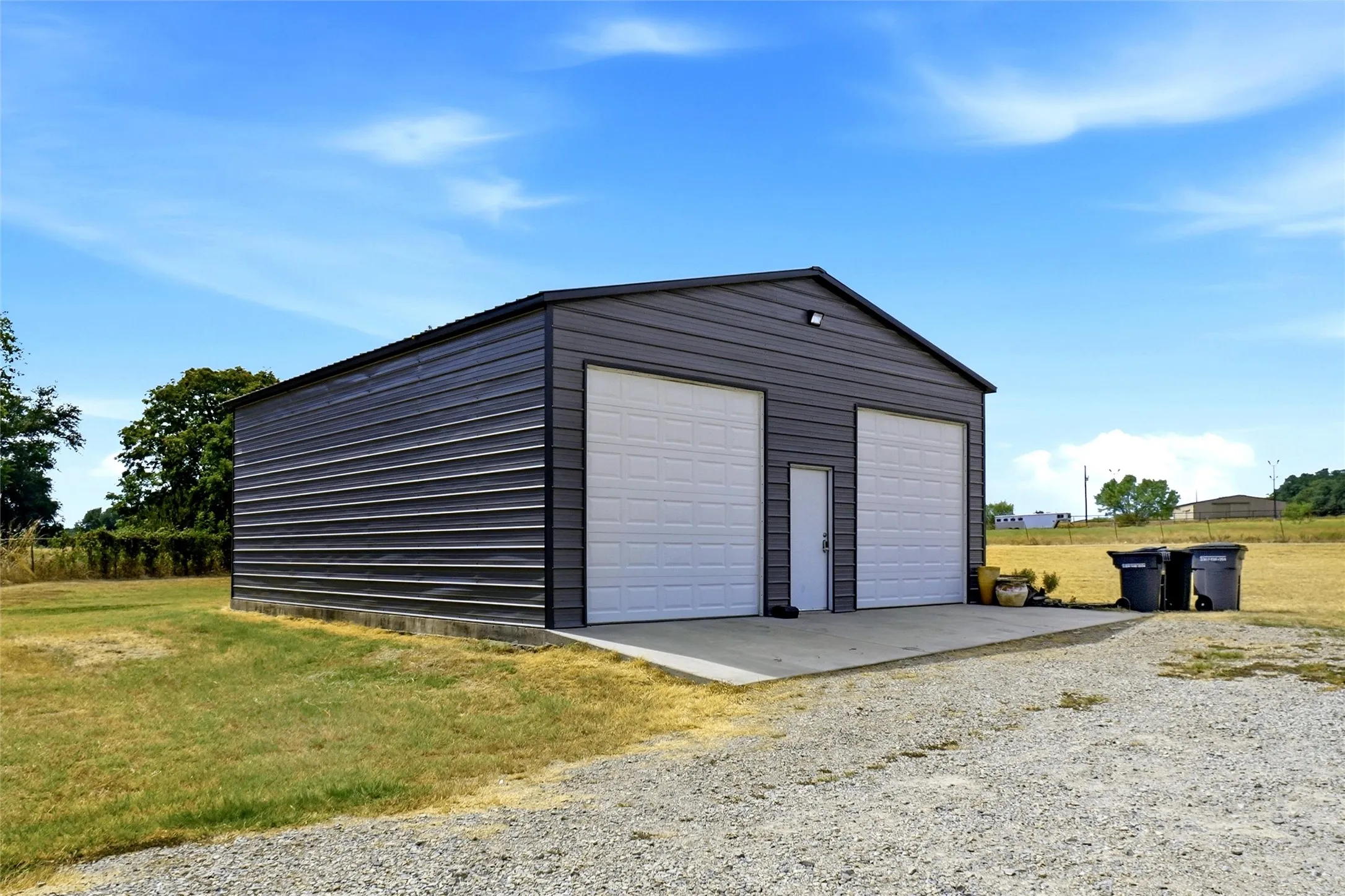 Two-bay metal structure with a gravel driveway, concrete pad, and exterior lighting