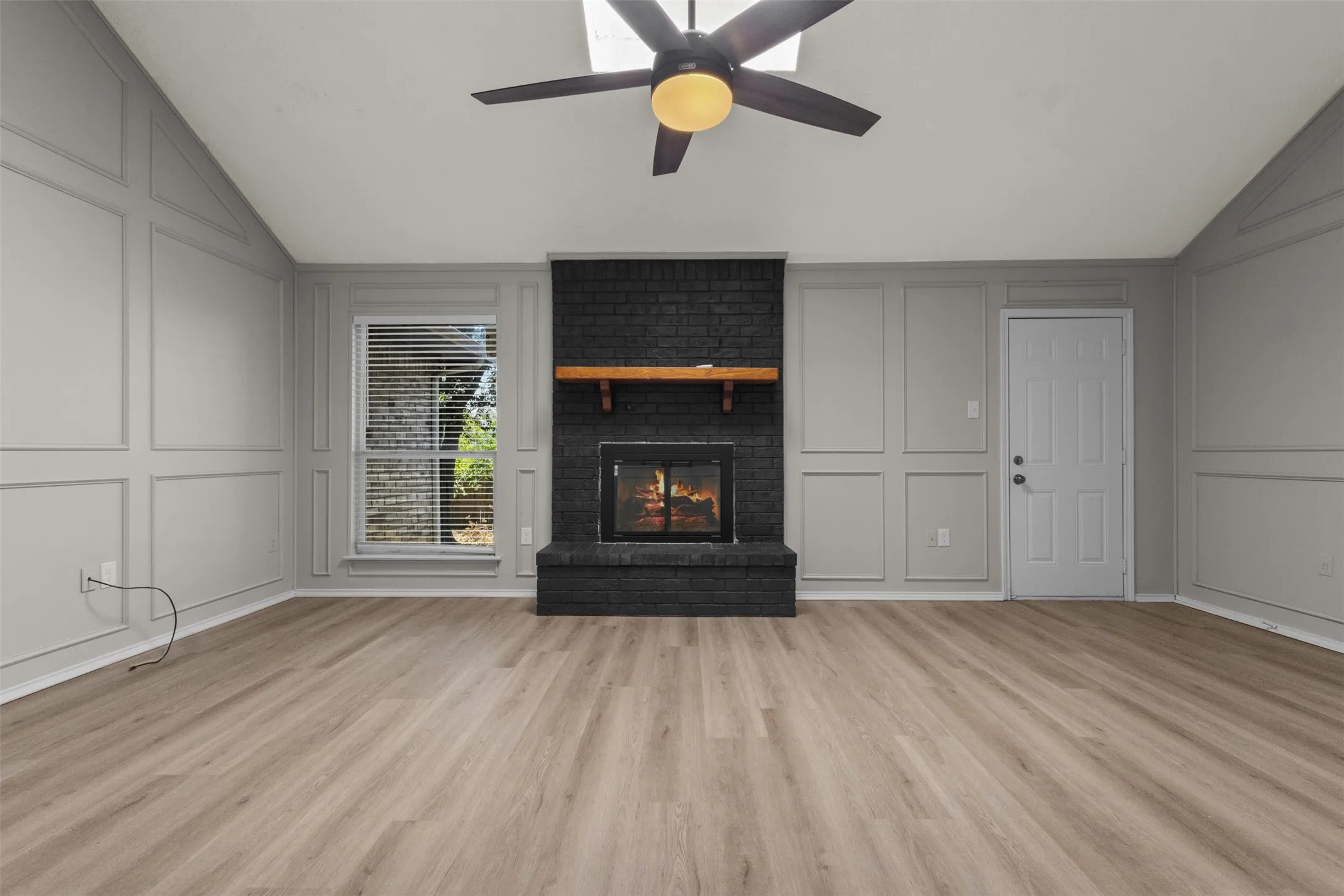 Wood-finish flooring, accent wall paneling, vaulted ceiling, black brick fireplace with wood mantel, and a ceiling fan with integrated lighting