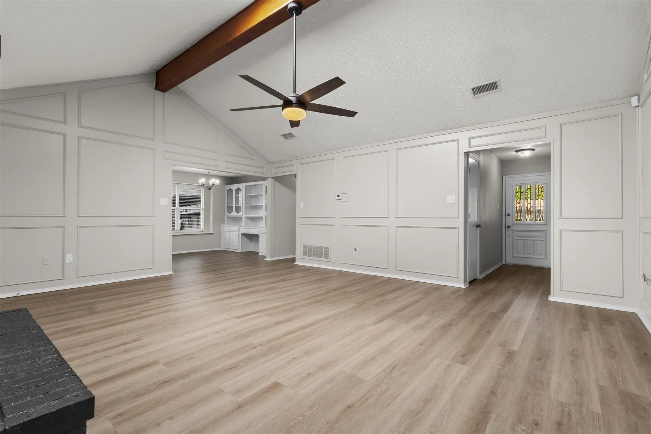 Vaulted ceiling with exposed wood beam, wood-finish flooring, decorative wall paneling, ceiling fan with integrated lighting, and built-in cabinetry with desk space