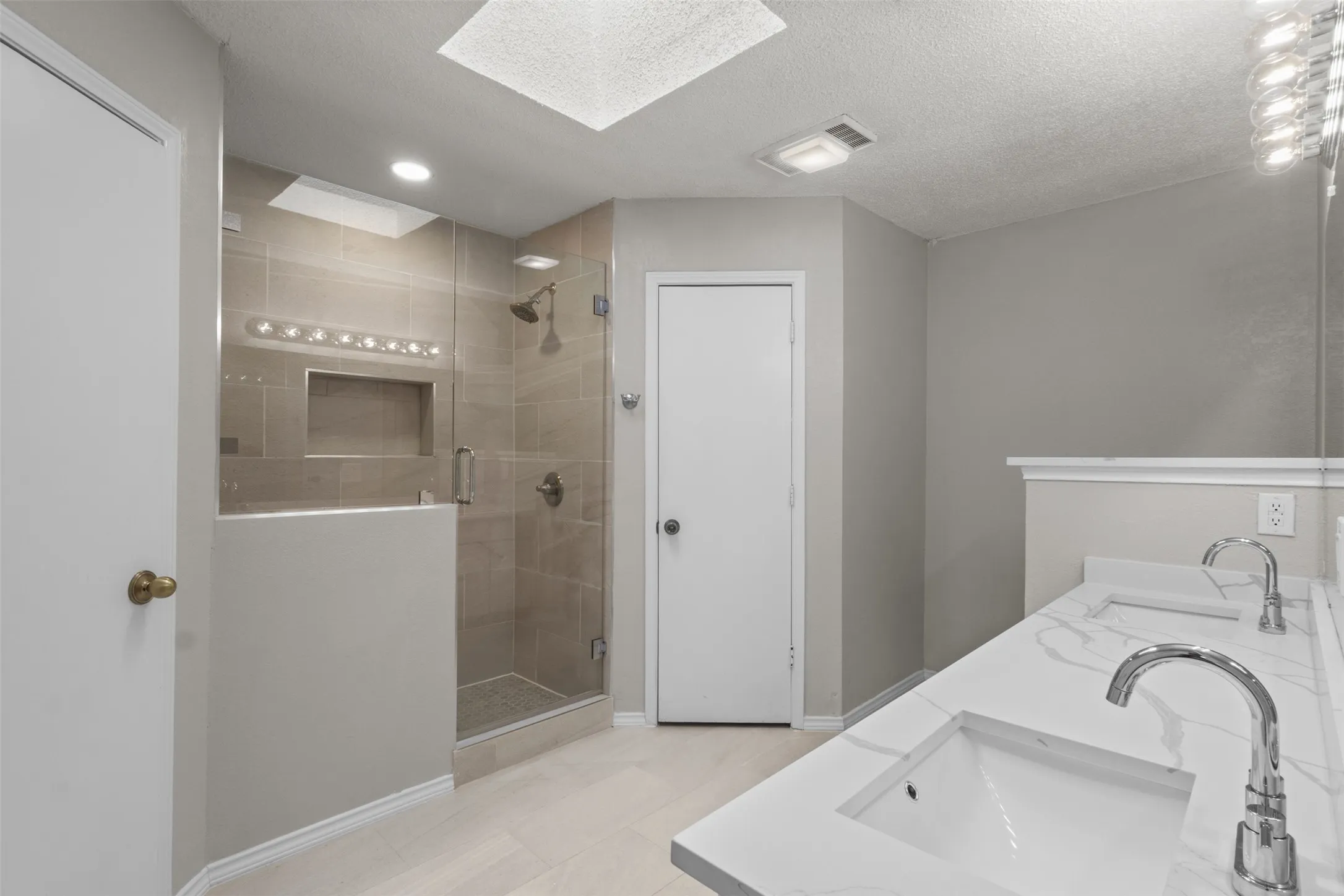 Dual vanity with integrated sinks, expansive shower with glass enclosure, tiled shower walls with recessed niche, overhead skylight, and wood-finish flooring