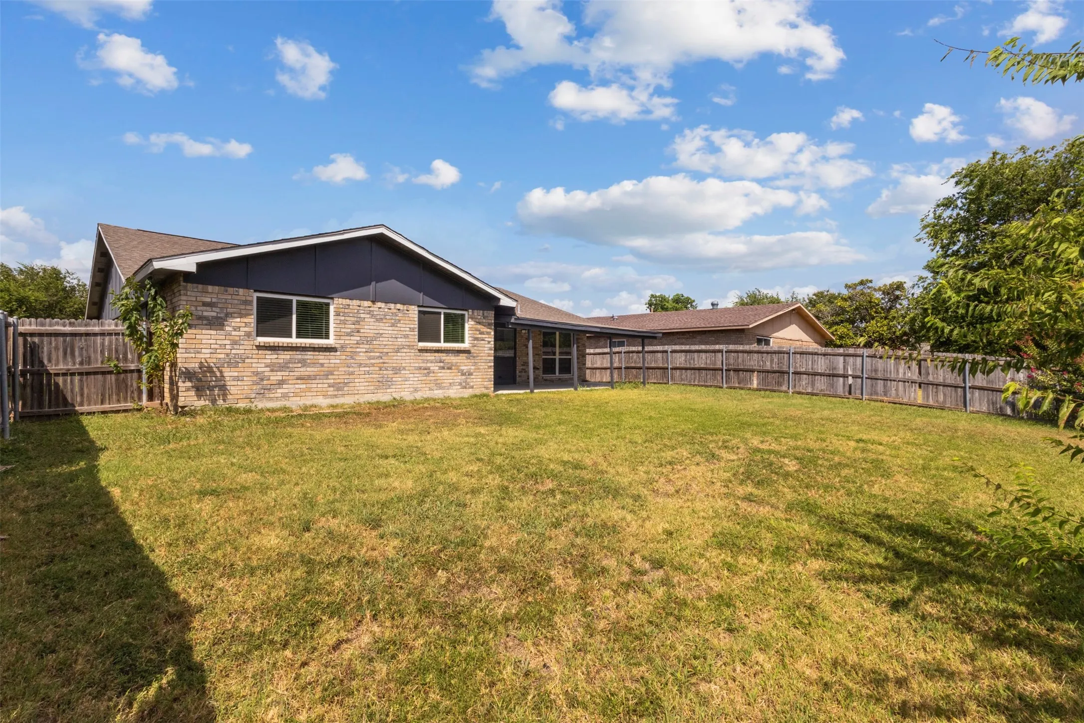Expansive fenced backyard with a brick and wood exterior, featuring multiple windows and a covered patio