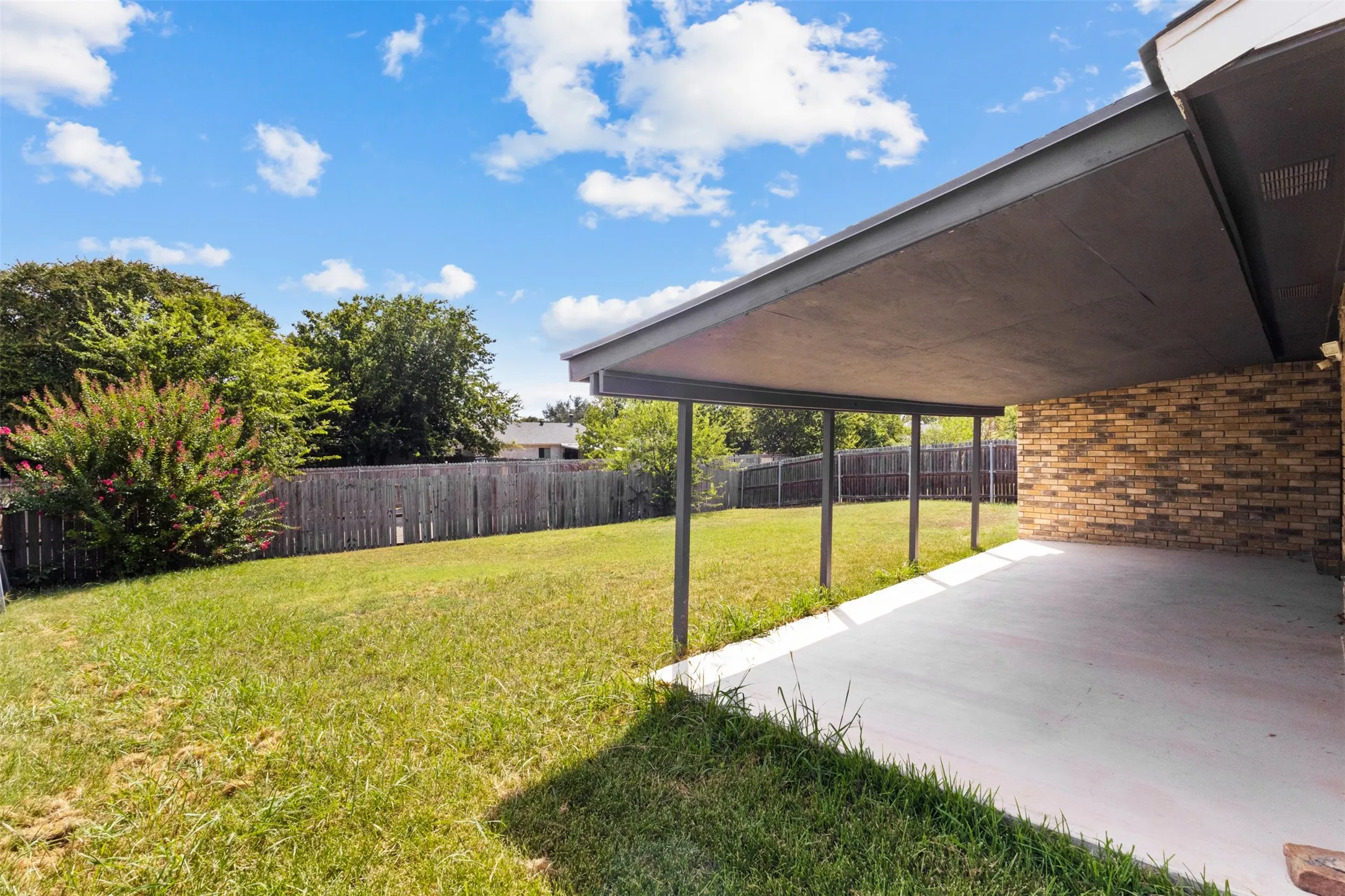 Covered patio with a concrete slab, brick exterior wall, and surrounding lawn with mature trees and a wooden fence