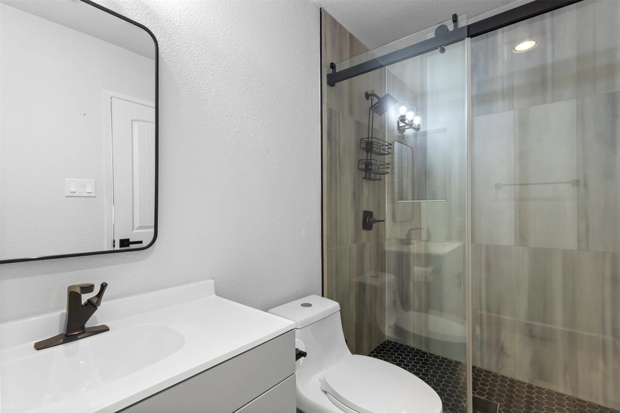 Modern bathroom featuring a single vanity with a white countertop, a rectangular mirror with a black frame, a toilet, a glass-enclosed shower with dark honeycomb tile flooring, and wood-finish wall tile