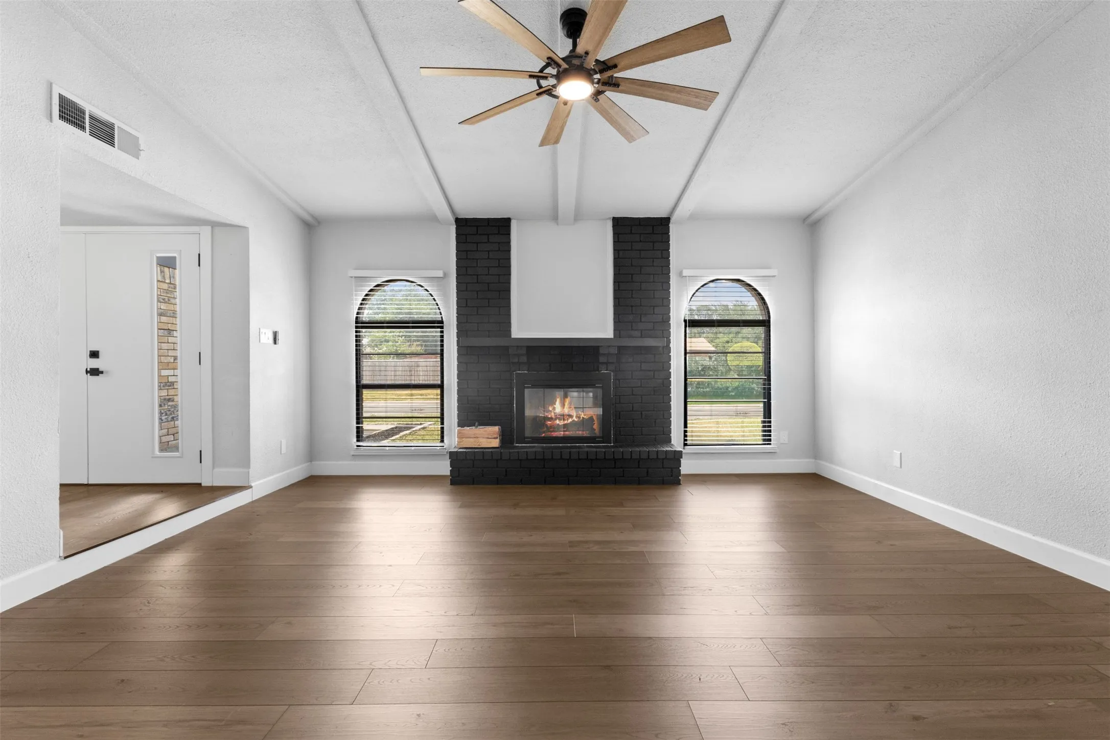 Wood-finish flooring, a ceiling fan with wood-tone blades, a black brick fireplace, and arched windows with horizontal blinds