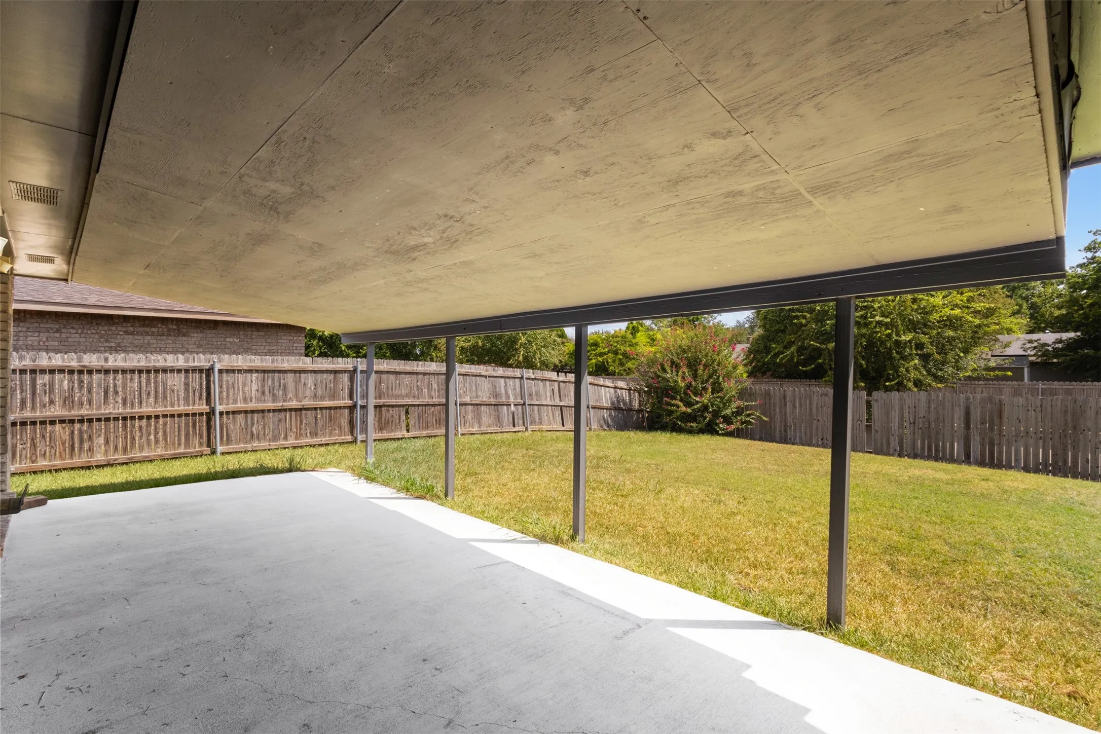 Expansive covered patio with concrete flooring, supported by metal posts, overlooking a fenced backyard with a grass lawn