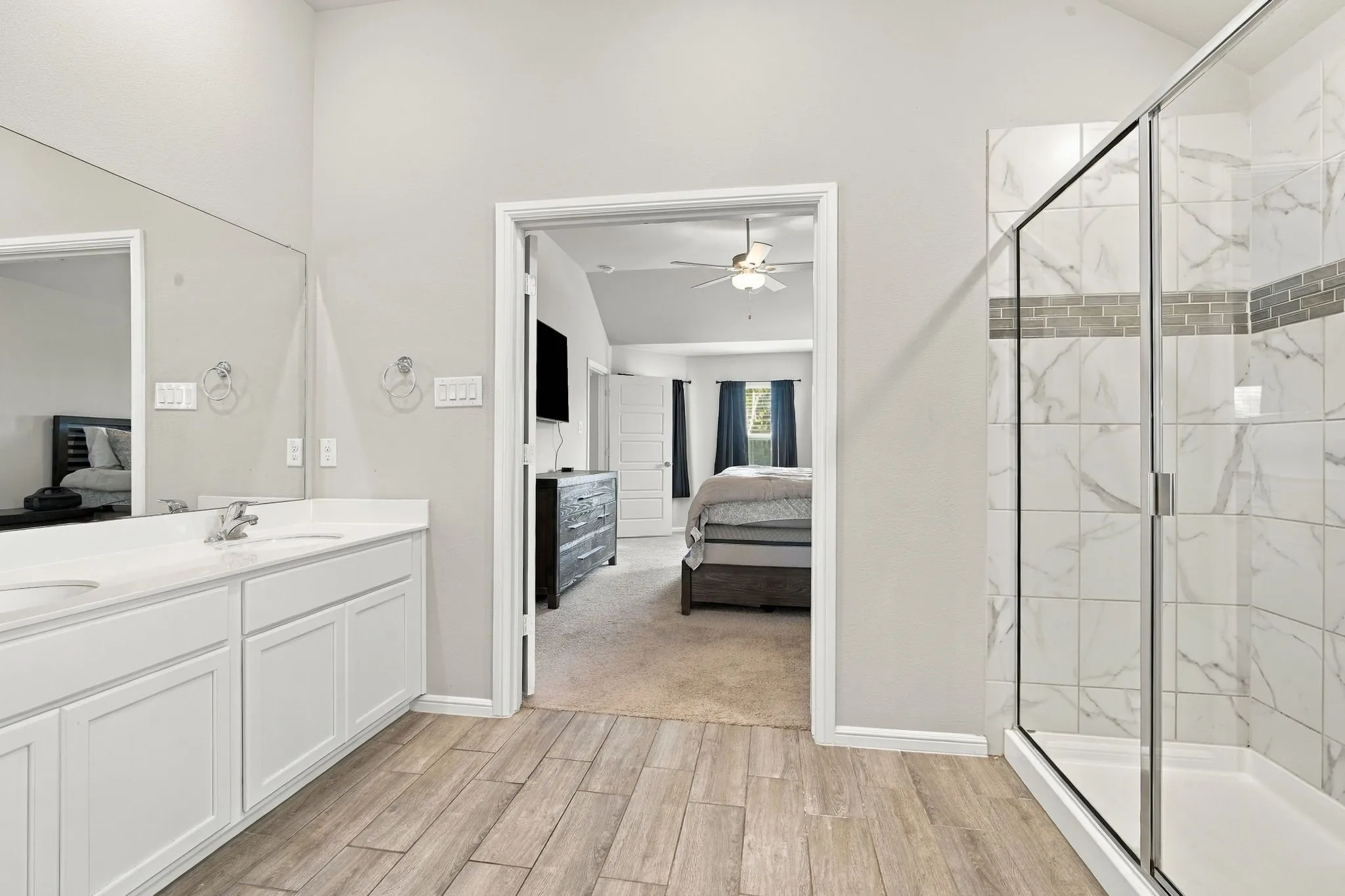 Dual vanity with undermount sinks, wood-finish flooring, glass-enclosed shower with tile surround, and vaulted ceiling