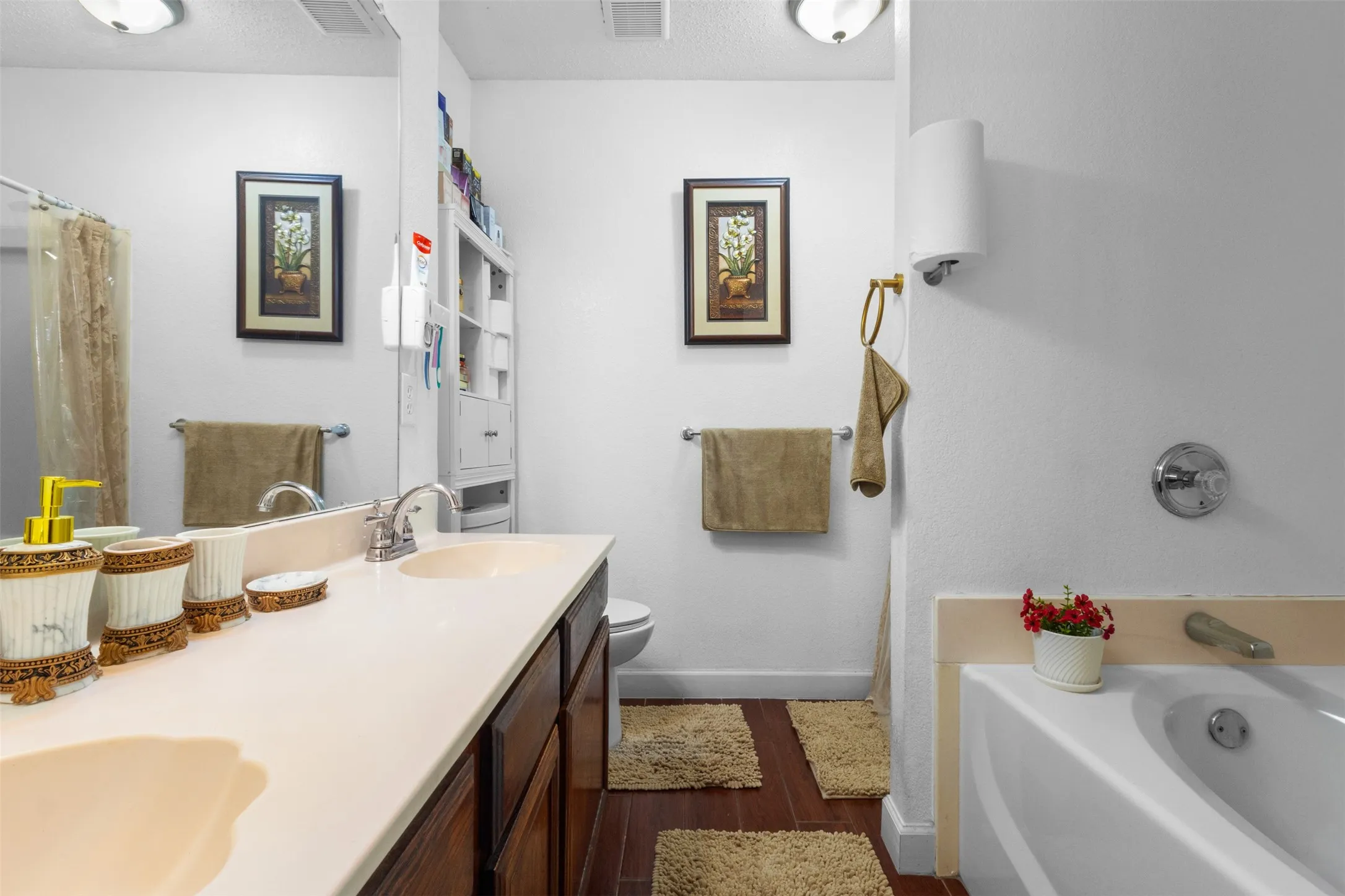 Double vanity with integrated sinks, rich wood-finish cabinetry, built-in linen storage, wood-finish flooring, and a full-size bathtub