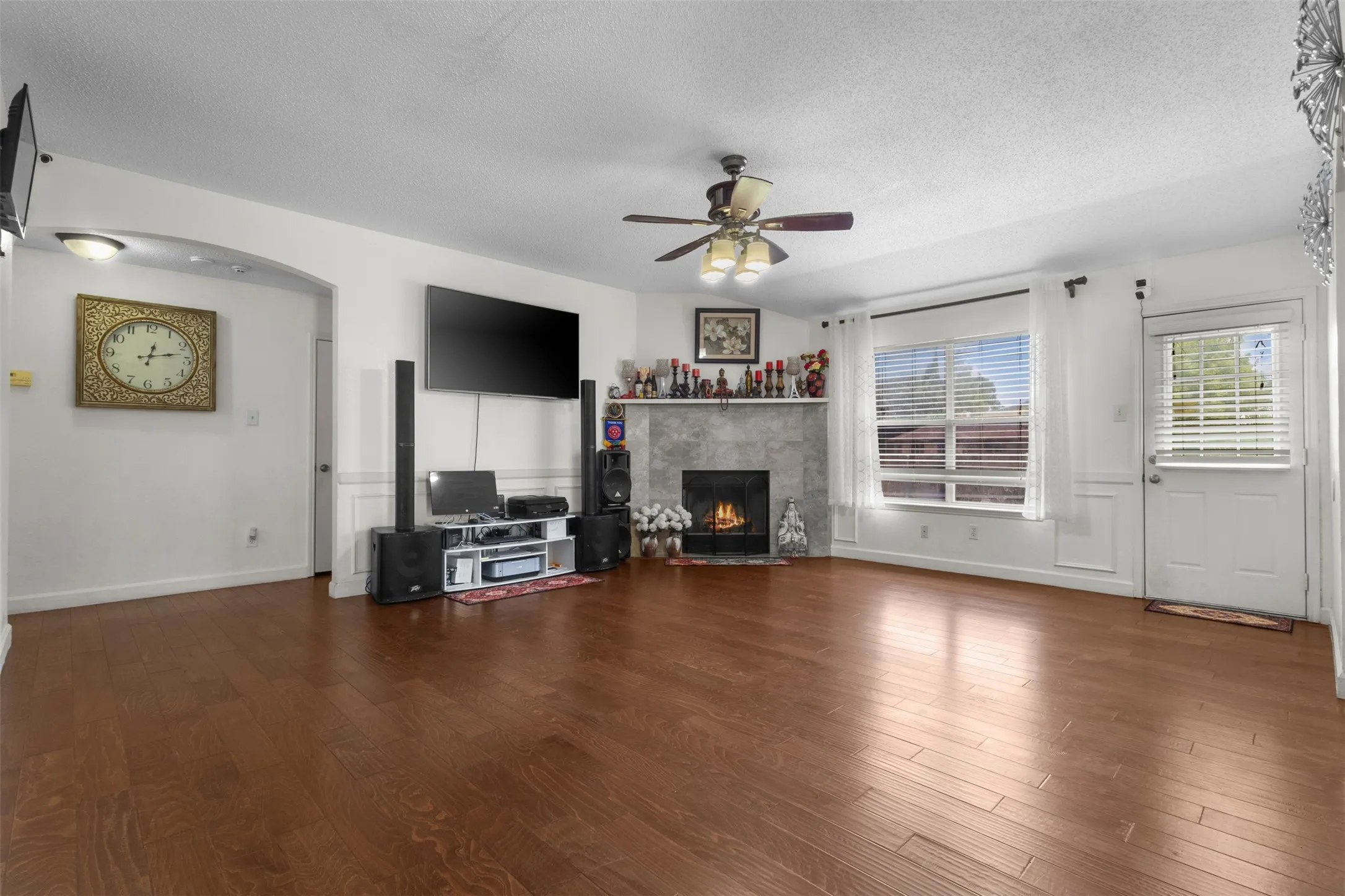 Wood-finish flooring, a stone-clad fireplace with mantel, a ceiling fan with integrated lighting, and a paneled exterior door