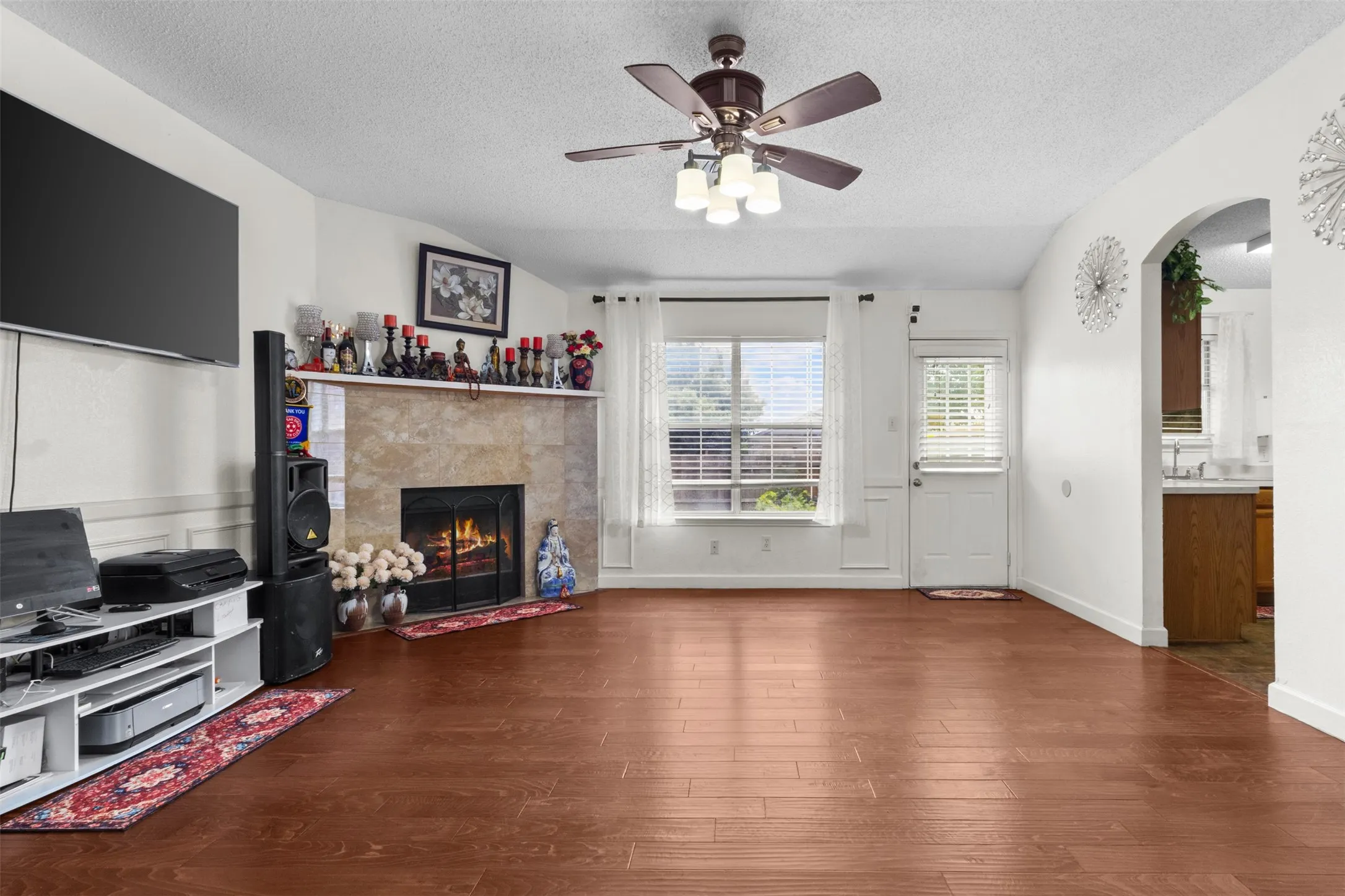 Living area featuring wood-finish flooring, a ceiling fan with integrated lighting, a fireplace with a stone surround, and an arched entry to an additional space