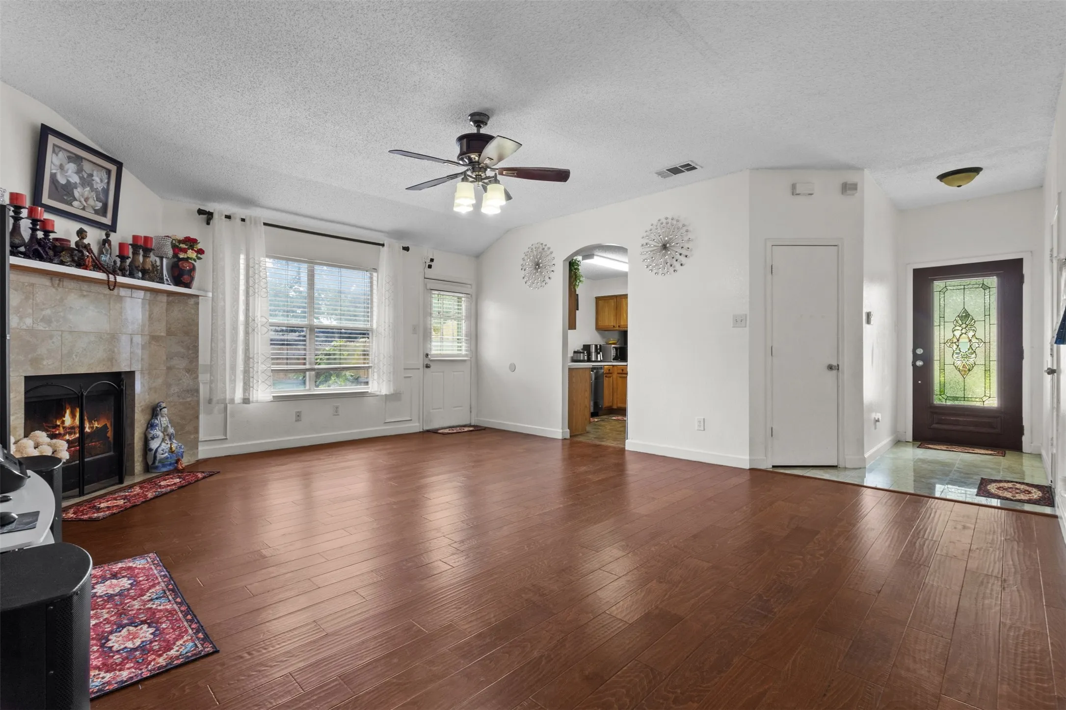 Wood-finish flooring throughout, a tiled fireplace with mantel, ceiling fan with integrated lighting, a glass-paneled entry door, and vaulted ceilings