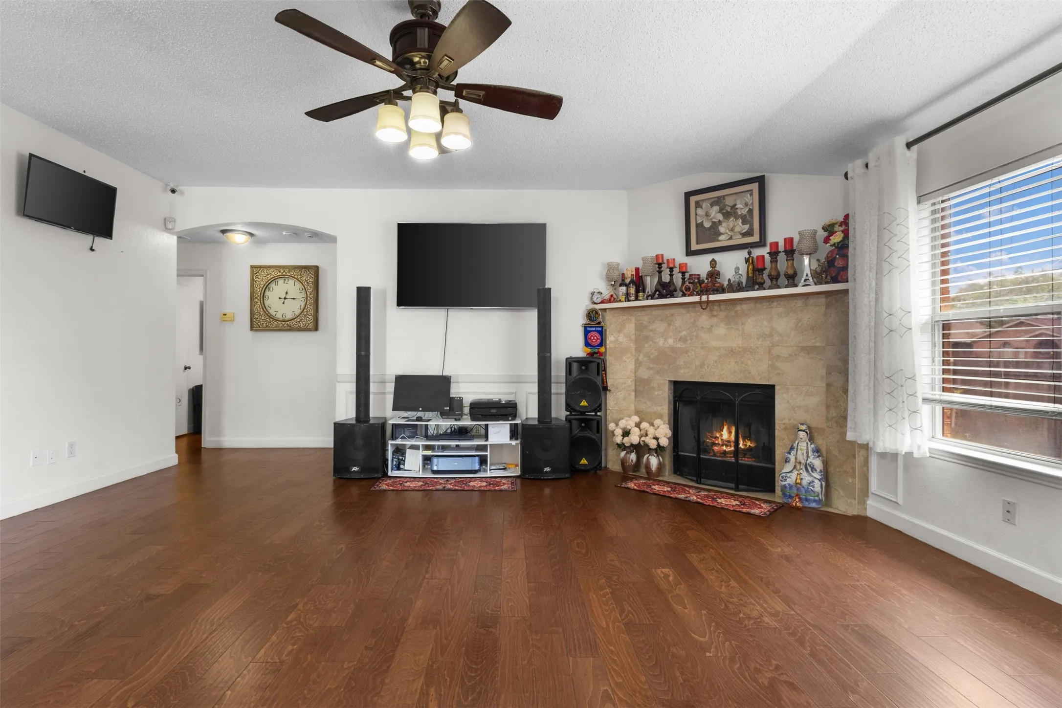Warm living space featuring wood-finish flooring, a ceiling fan with integrated lighting, a stone-clad fireplace with a hearth, and an arched doorway
