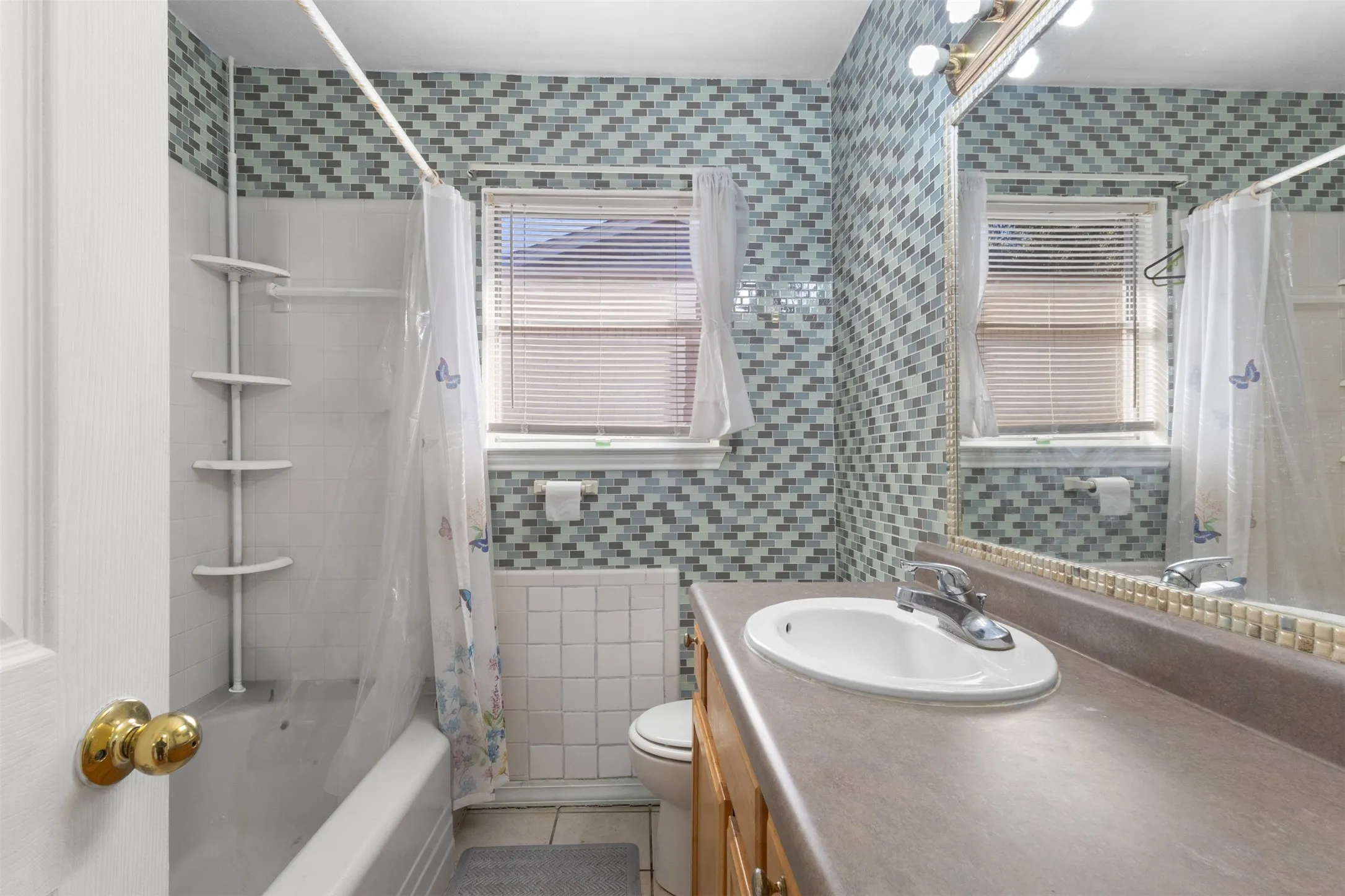 Bathroom featuring a full vanity with an integrated sink, a wall-mounted mirror with overhead lighting, a bathtub with a shower, and a mosaic tile wall
