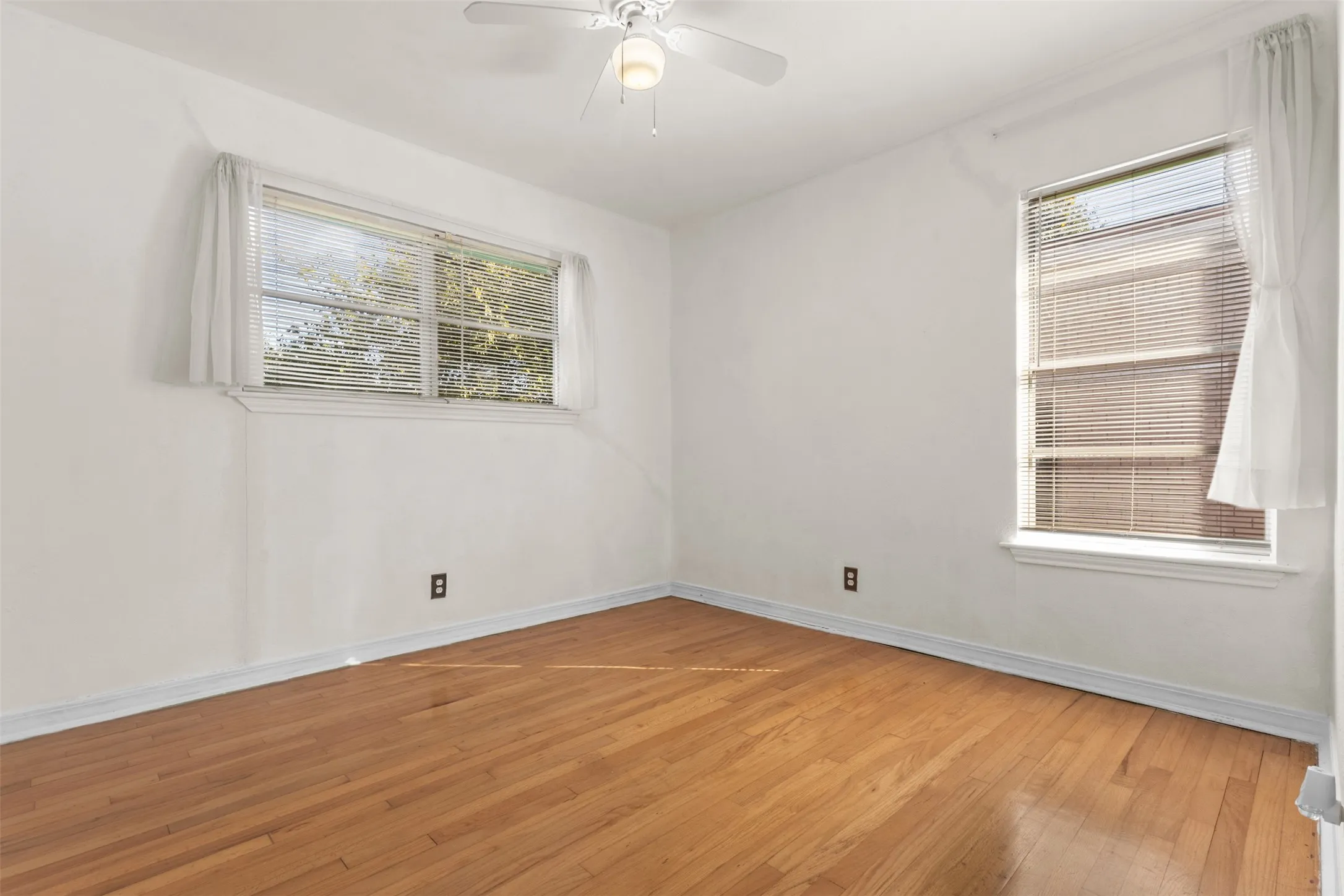 Wood-finish flooring, white walls, two windows with blinds, and a ceiling fan with integrated lighting