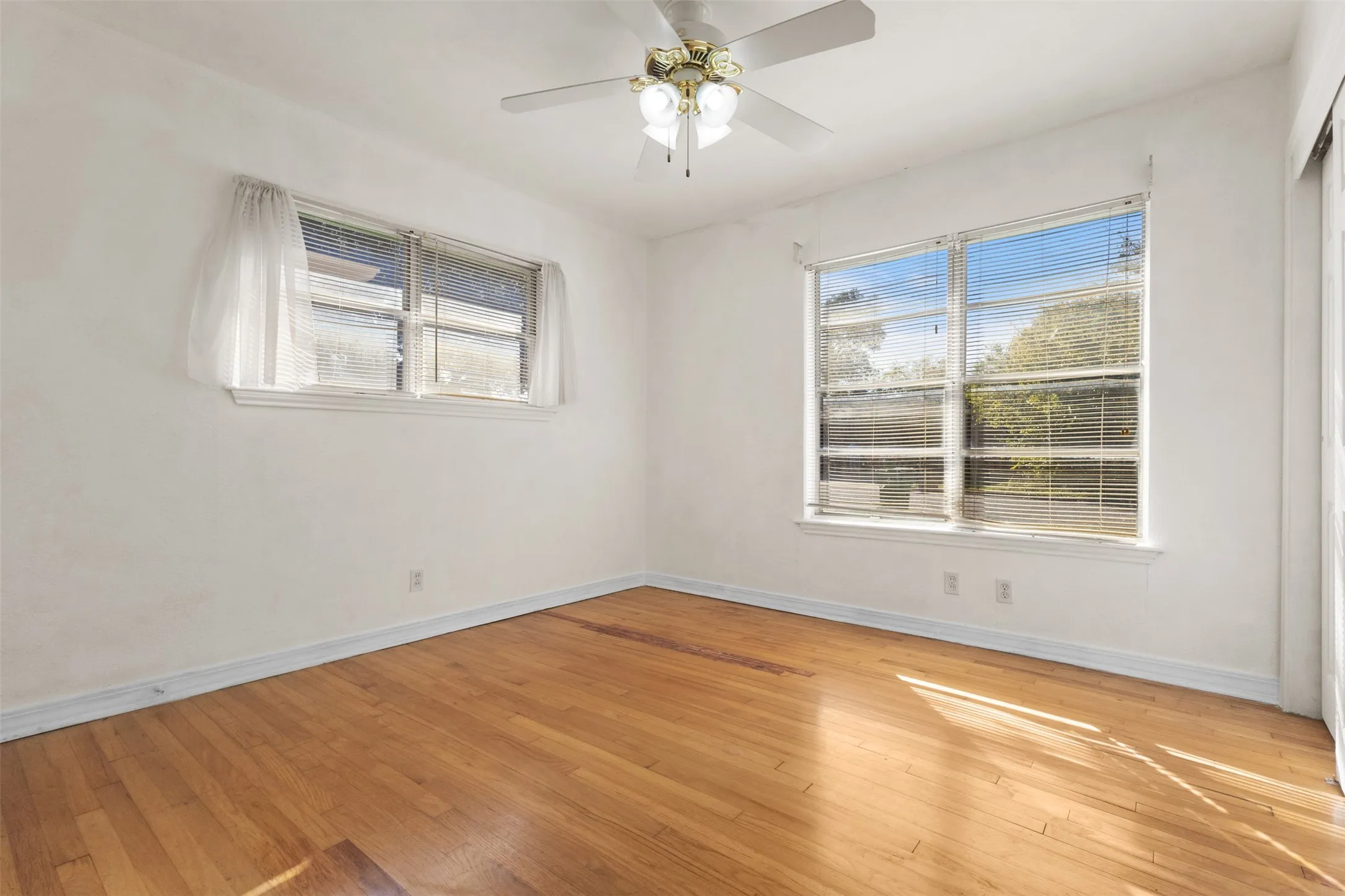 Hardwood flooring, white walls, two windows with blinds, and a ceiling fan with integrated lighting