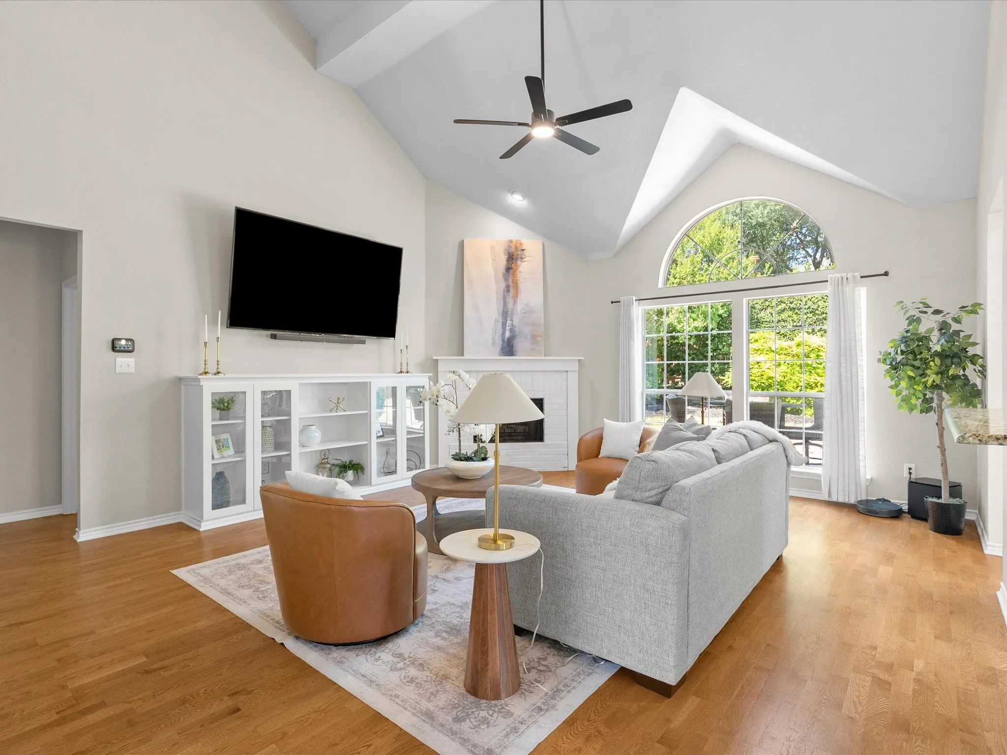 Vaulted ceiling, wood-finish flooring, white built-in cabinetry, fireplace with white mantel, and arched window with exterior landscaping