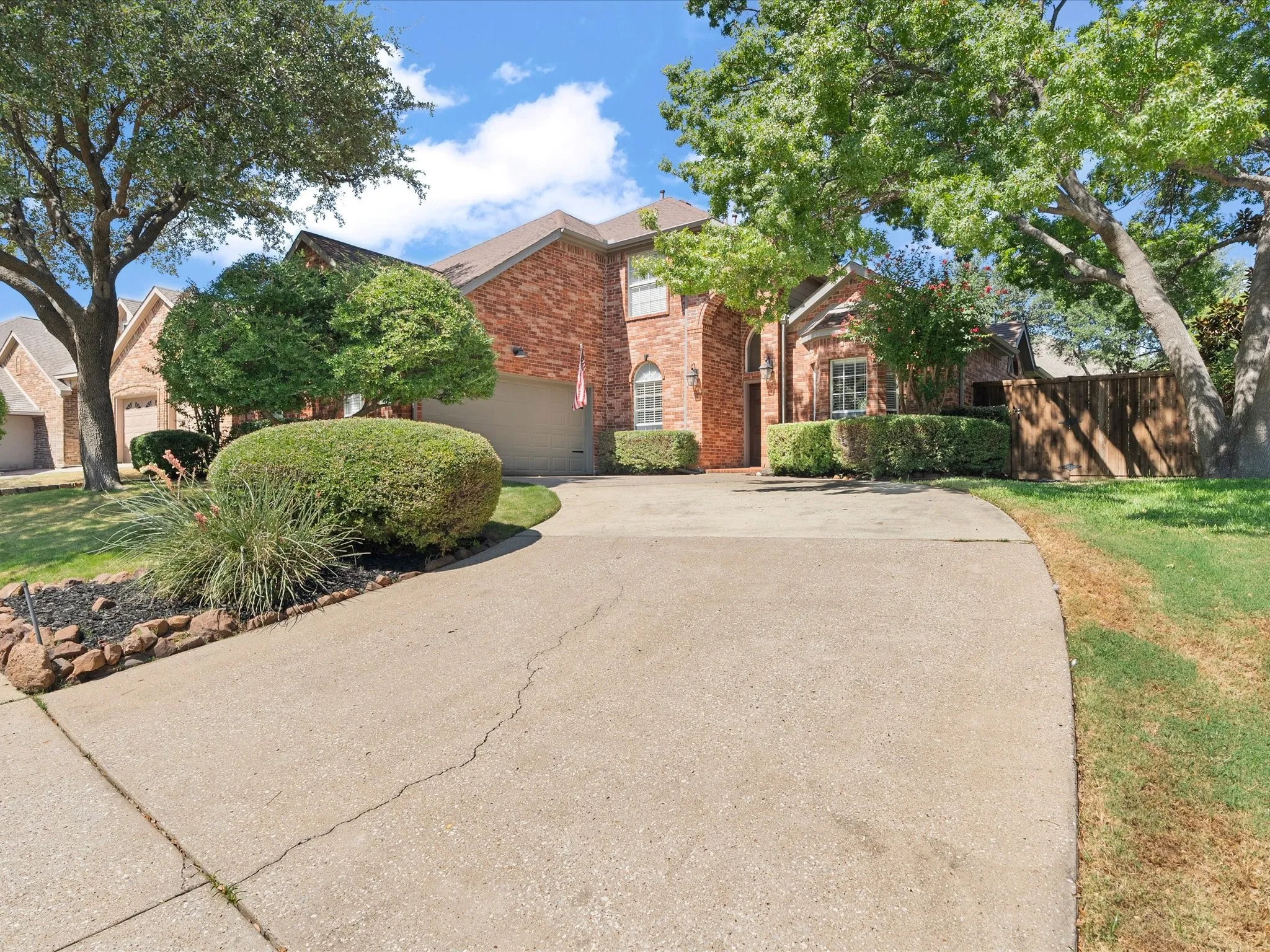 Brick exterior construction with a two-car garage, mature trees, manicured shrubs, and a concrete driveway