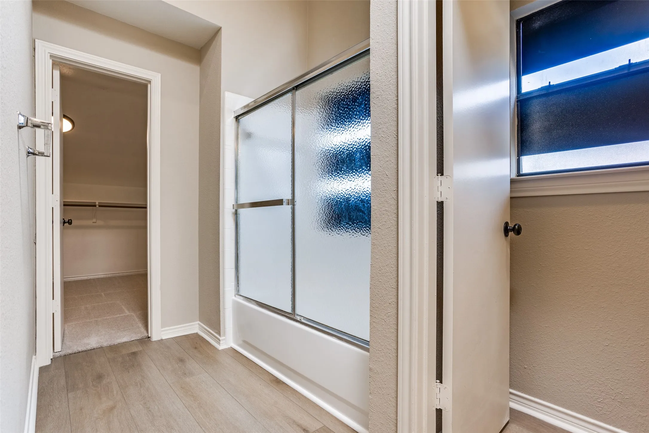 Wood-finish flooring, full-size bathtub with sliding privacy glass doors, and a window with privacy film