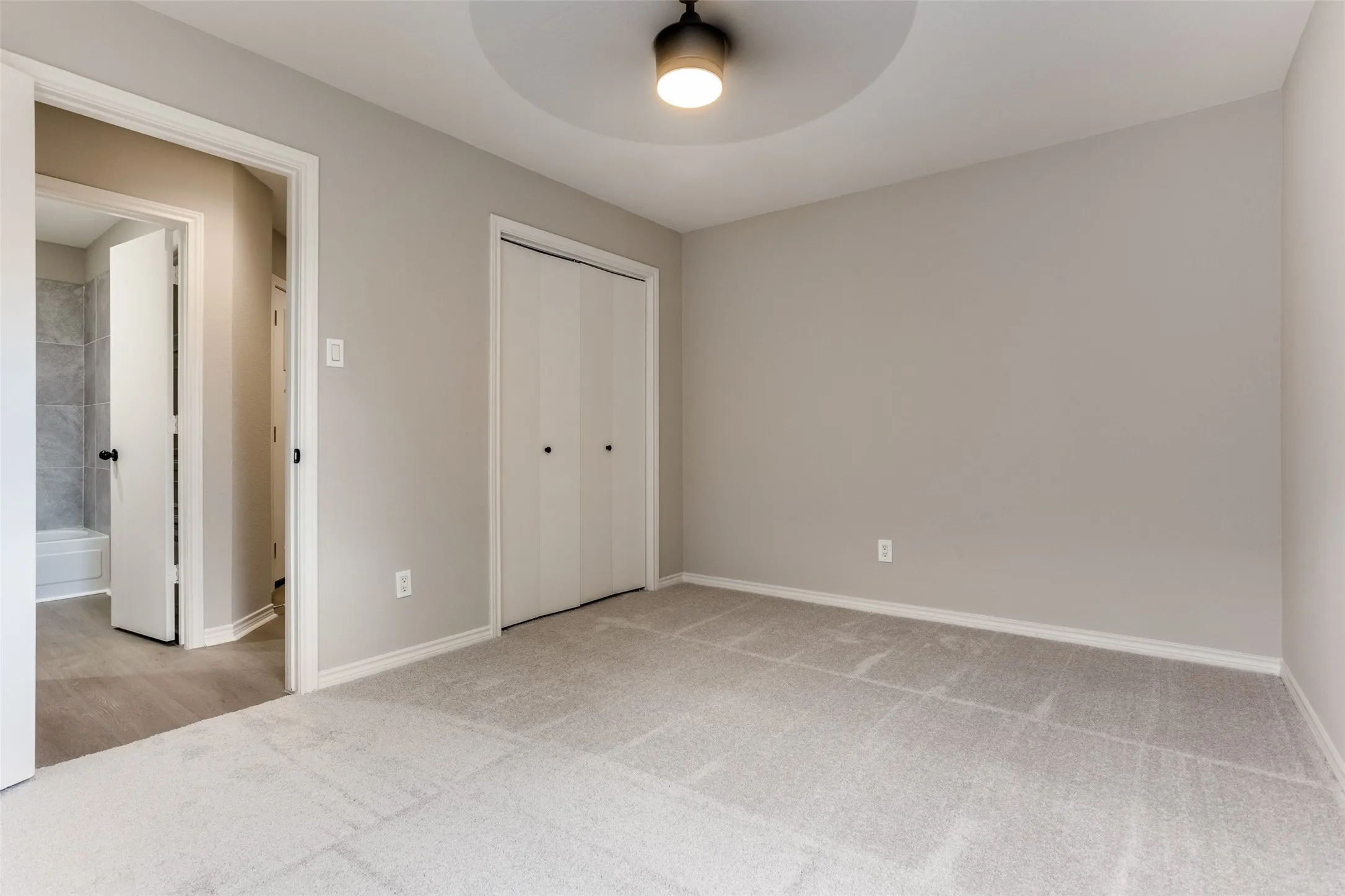 Light gray wall paint, carpeting, ceiling light fixture with a decorative ceiling medallion, double-door closet with black hardware, and an en-suite bathroom with a tiled tub surround