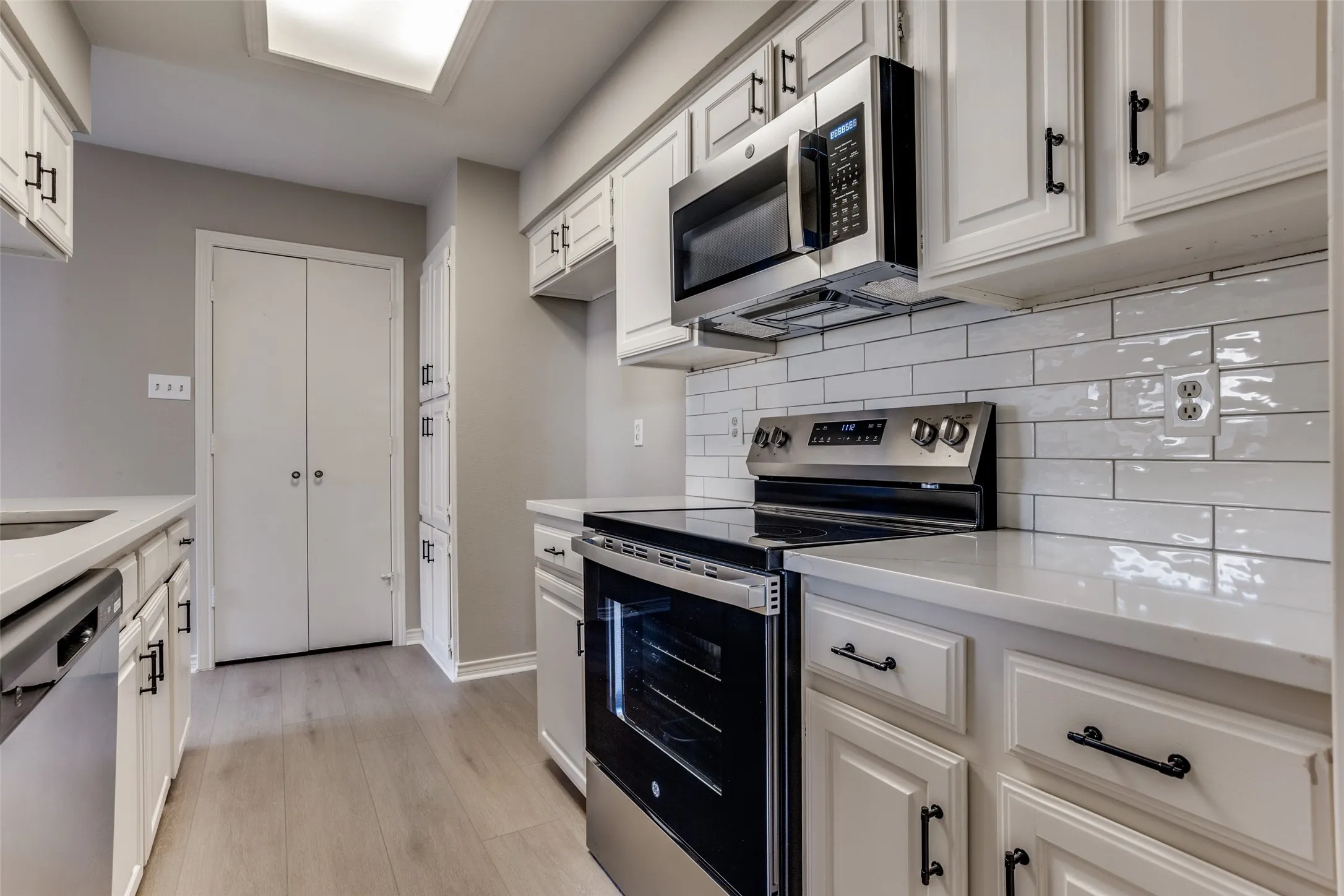 White cabinetry with black hardware, white subway tile backsplash, stainless steel appliances, light-toned wood-finish flooring, and light-toned countertops