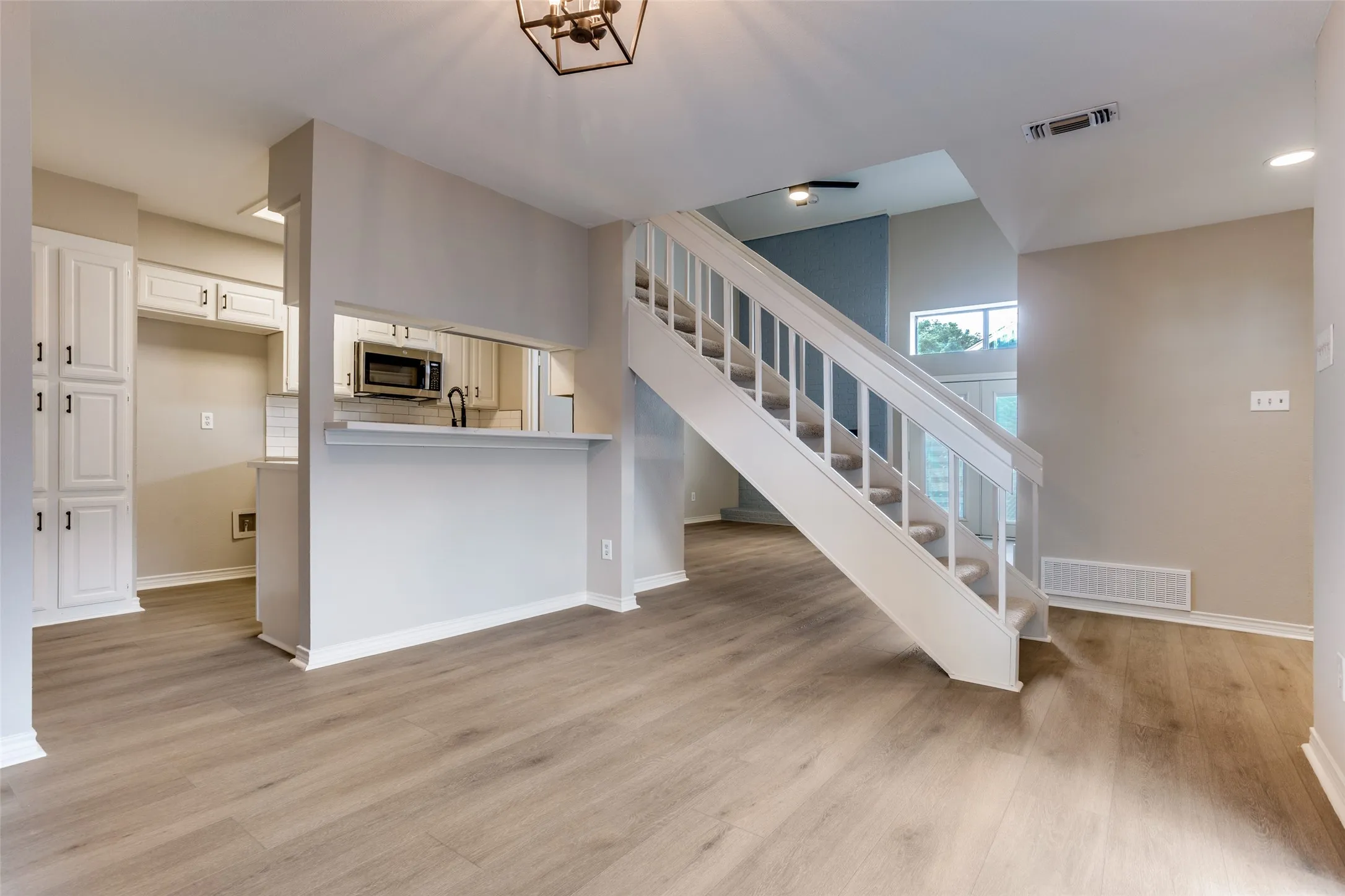 Wood-finish flooring throughout, white kitchen cabinetry with black hardware, pass-through kitchen window, white staircase with carpeting, and a modern ceiling light fixture