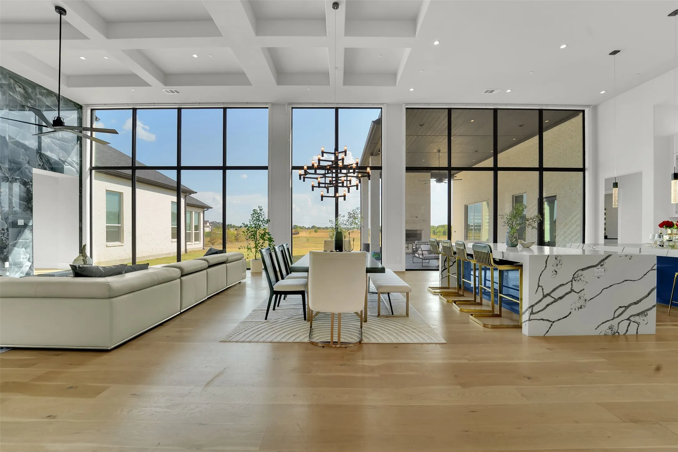 Expansive floor-to-ceiling windows, coffered ceiling, light wood-finish flooring, large kitchen island with integrated seating, and a striking accent wall