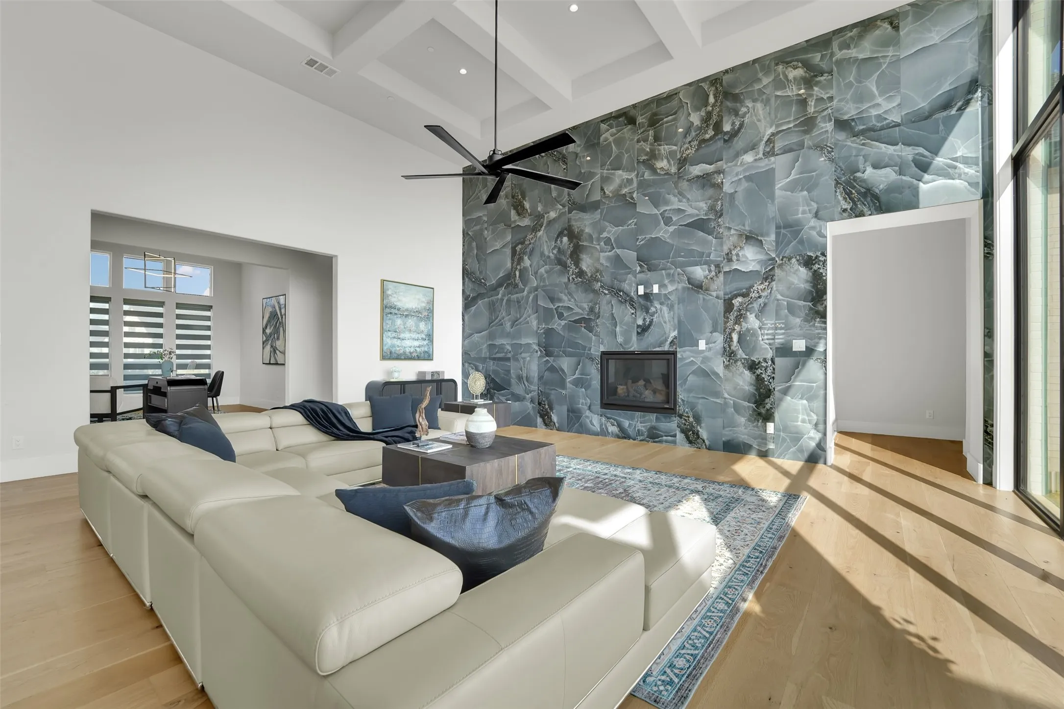 High ceilings with coffered details, expansive stone-finish accent wall with integrated fireplace, light wood-finish flooring, and a large multi-pane window