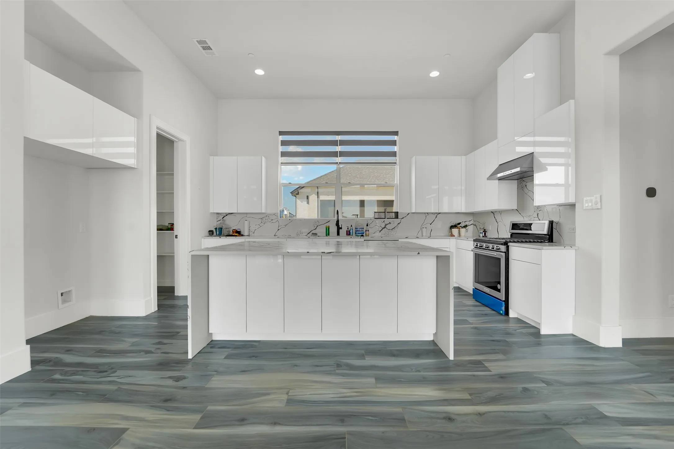 Expansive kitchen featuring wood-finish flooring, a central island with storage, white high-gloss cabinetry, and a gas range with an overhead vent hood