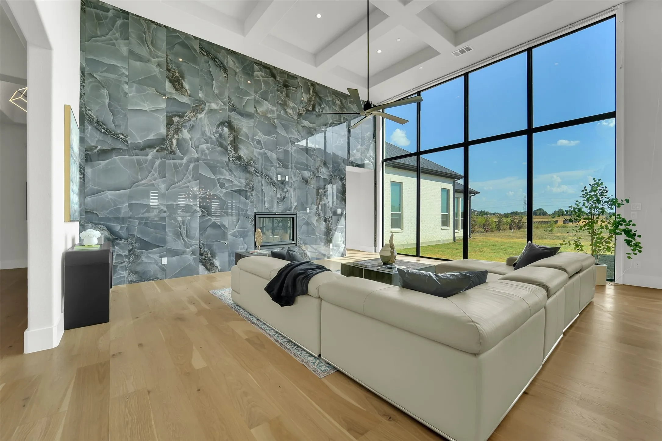 Soaring ceilings with coffered details, a full-height stone-clad fireplace, expansive floor-to-ceiling windows, natural wood-finish flooring, and a contemporary ceiling fan