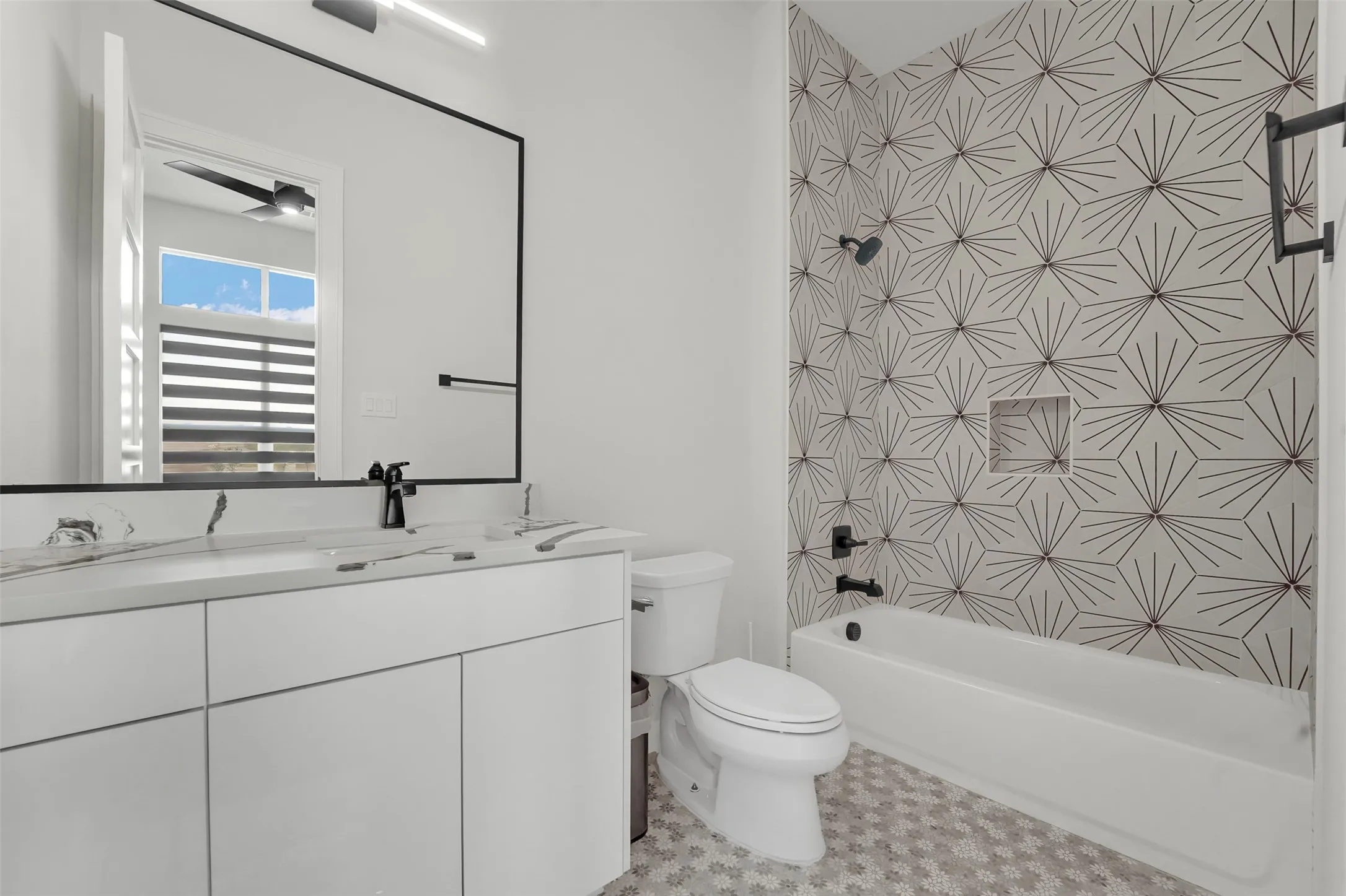 White double vanity with a marble-finish countertop, matte black fixtures, full-width mirror, patterned hexagonal tile accent wall, and a built-in shower niche