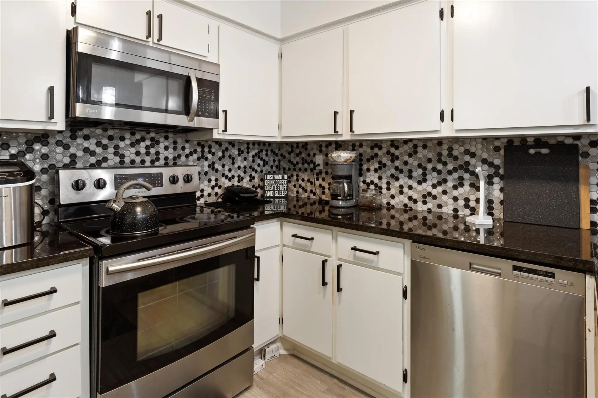 White cabinetry with matte black hardware, dark countertops, hexagonal mosaic tile backsplash, stainless steel appliances, and wood-finish flooring