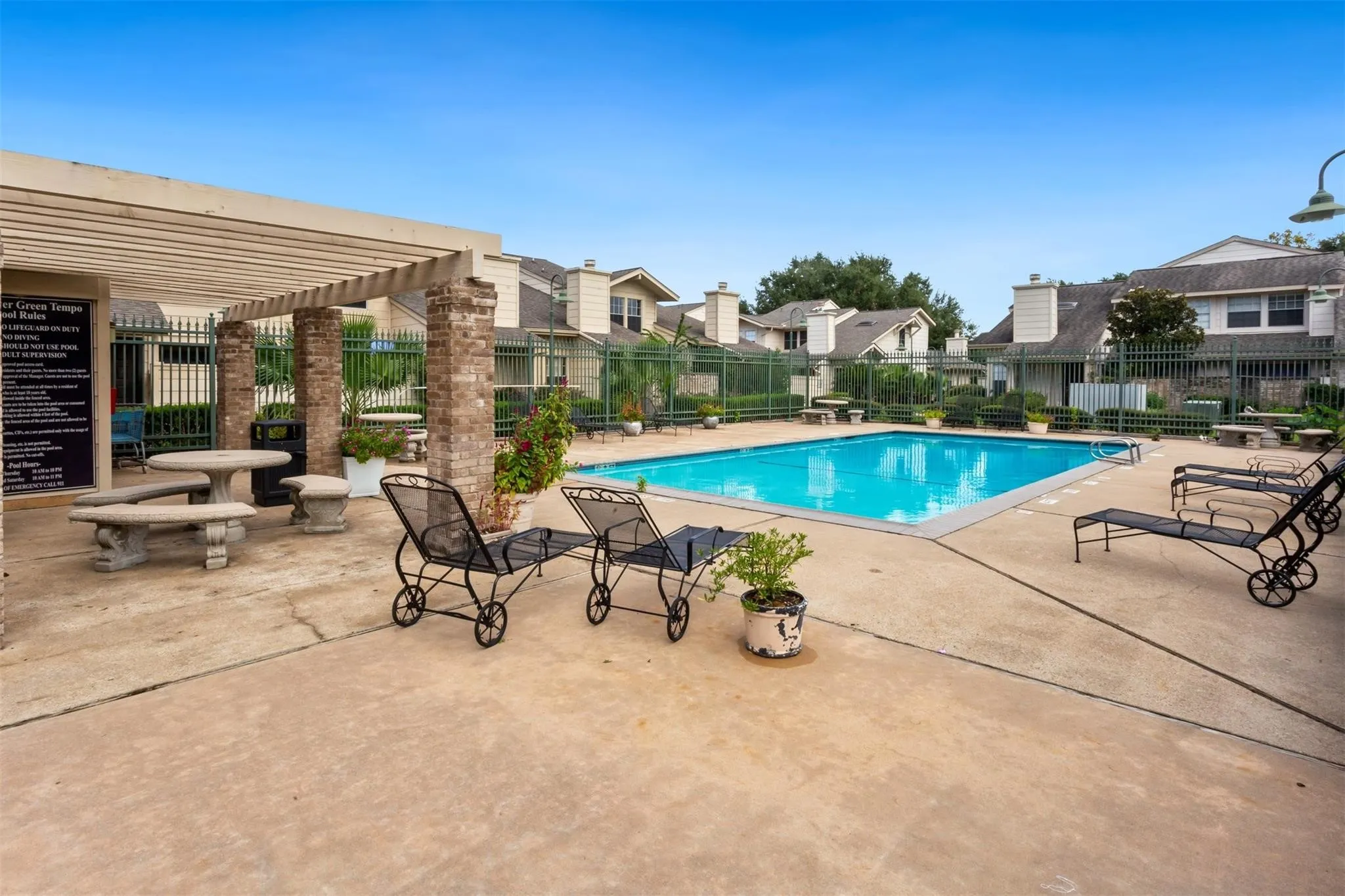 Community swimming pool with a surrounding concrete deck, shaded pergola with brick columns, and a perimeter fence