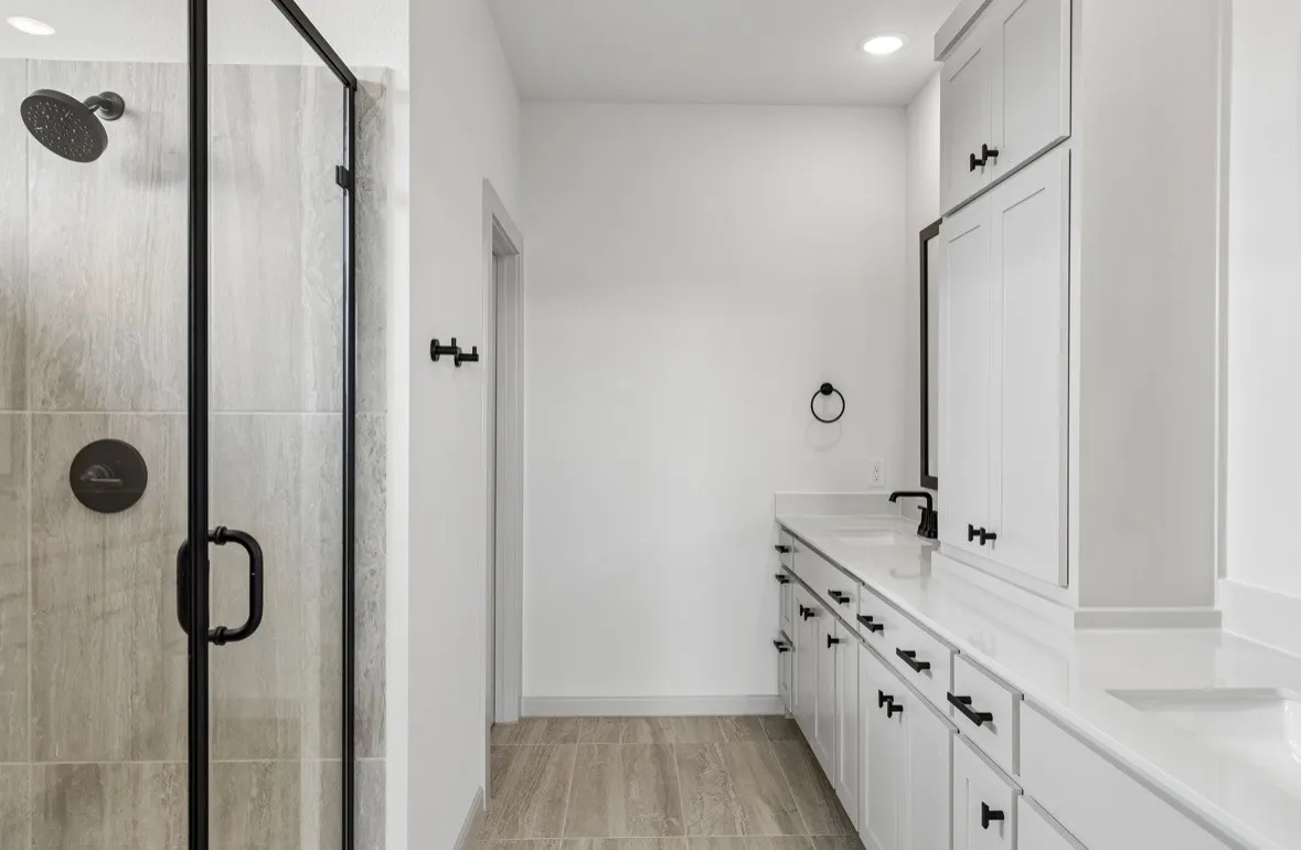 Walk-in shower with tile surround and dark bronze fixtures, dual vanity with white cabinetry, integrated sinks, white countertops, and wood-finish flooring