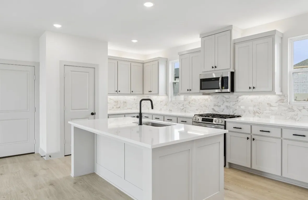 Kitchen featuring a central island with an integrated sink, light wood-finish flooring, white cabinetry with black hardware, stone countertops, and a tiled backsplash