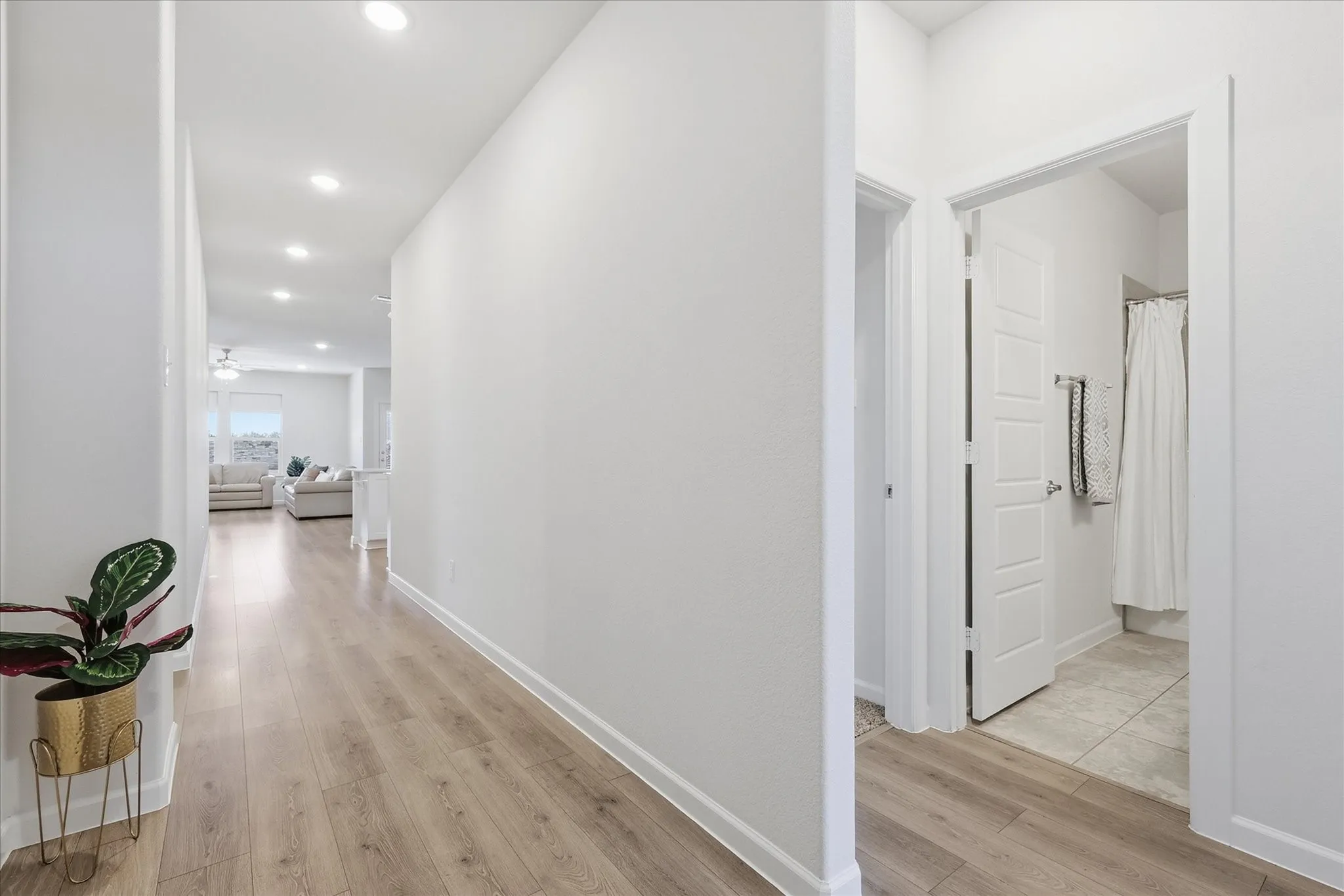 Recessed lighting illuminates the hallway with wood-finish flooring, leading to a bathroom featuring ceramic tile flooring and a shower with a white shower curtain