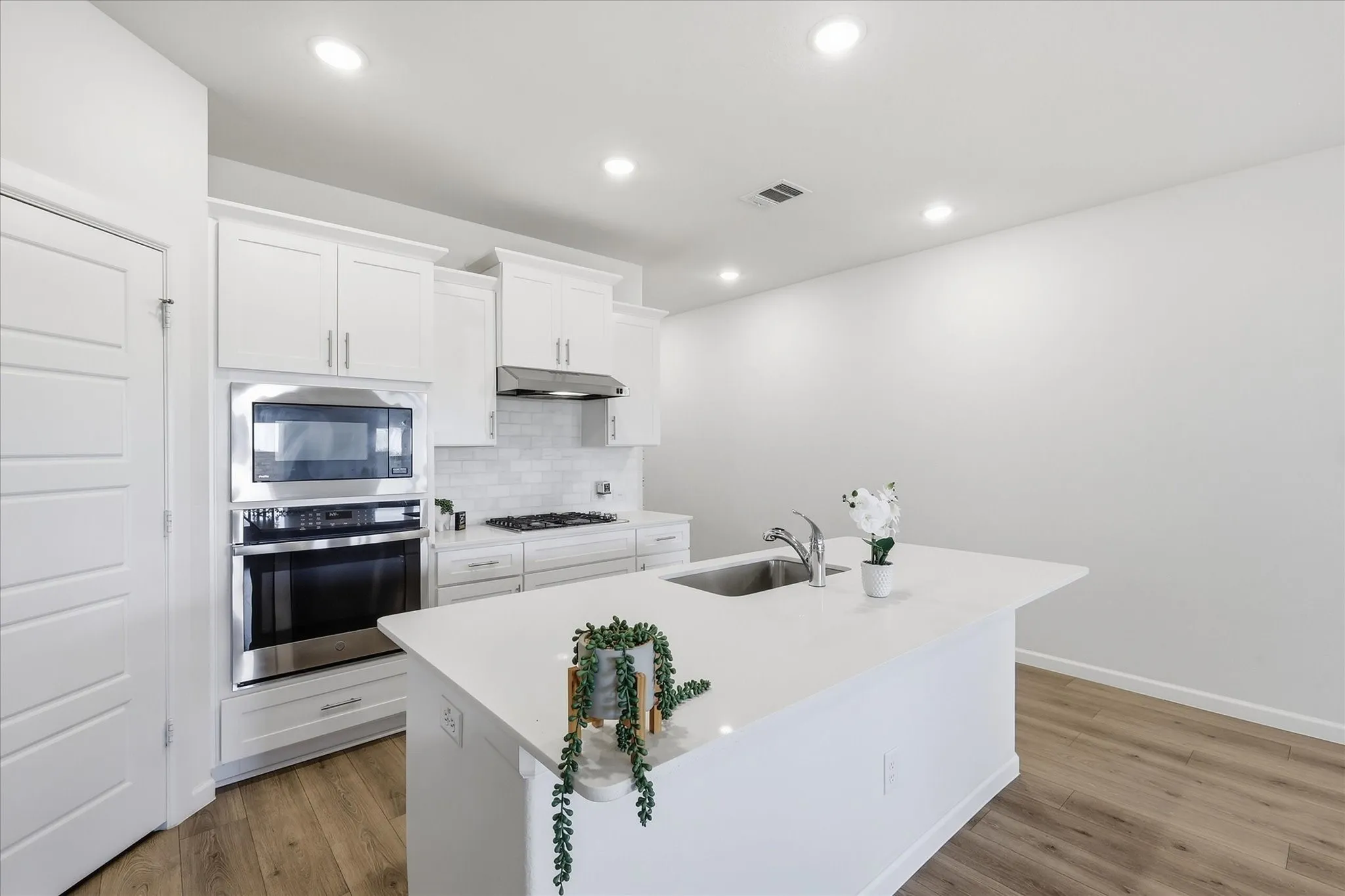White shaker cabinetry with crown molding, stainless steel built-in appliances, quartz countertops, subway tile backsplash, and wood-finish flooring