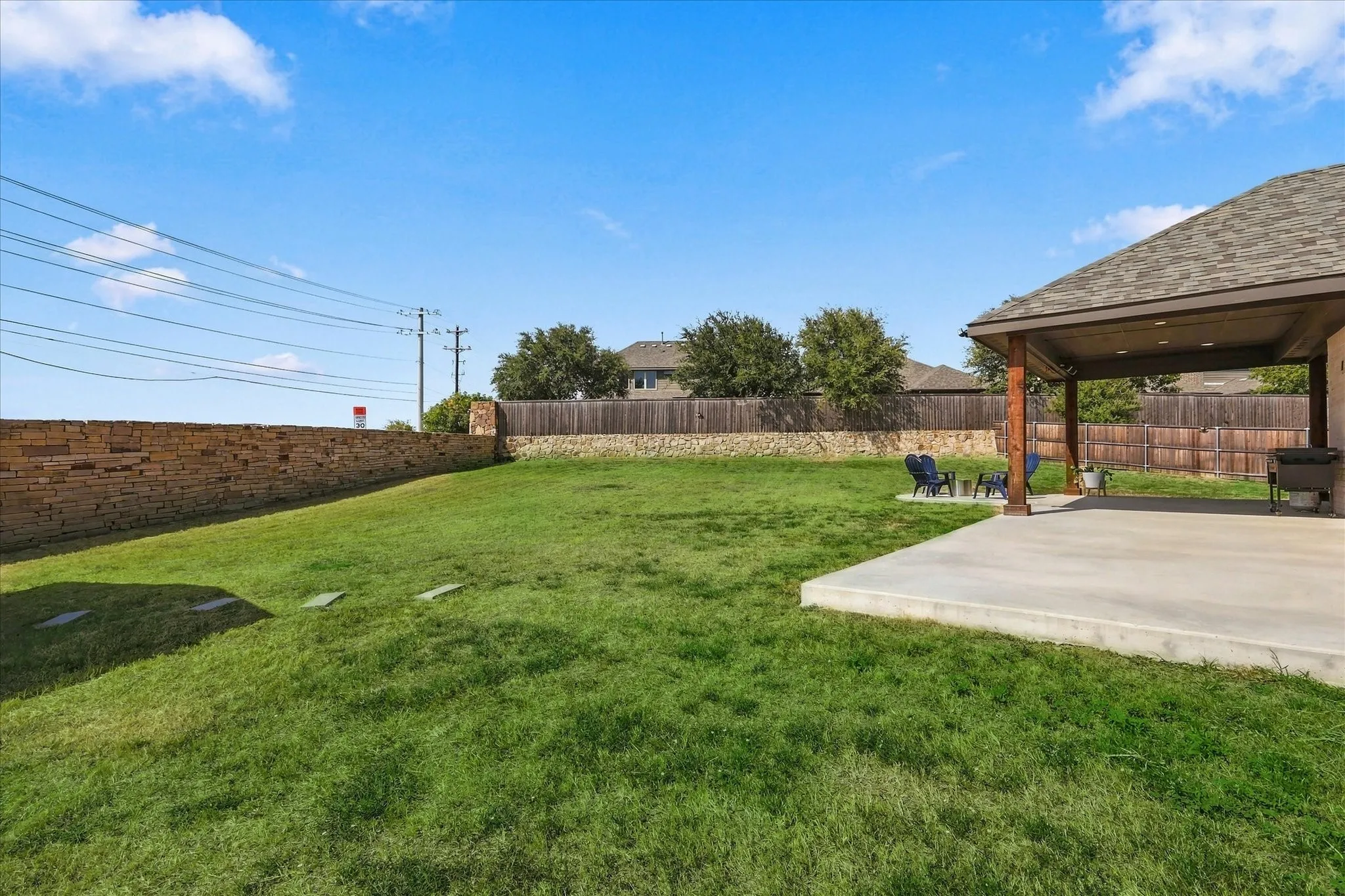 Expansive backyard featuring a concrete patio, covered outdoor living space with wood posts, and a stone retaining wall