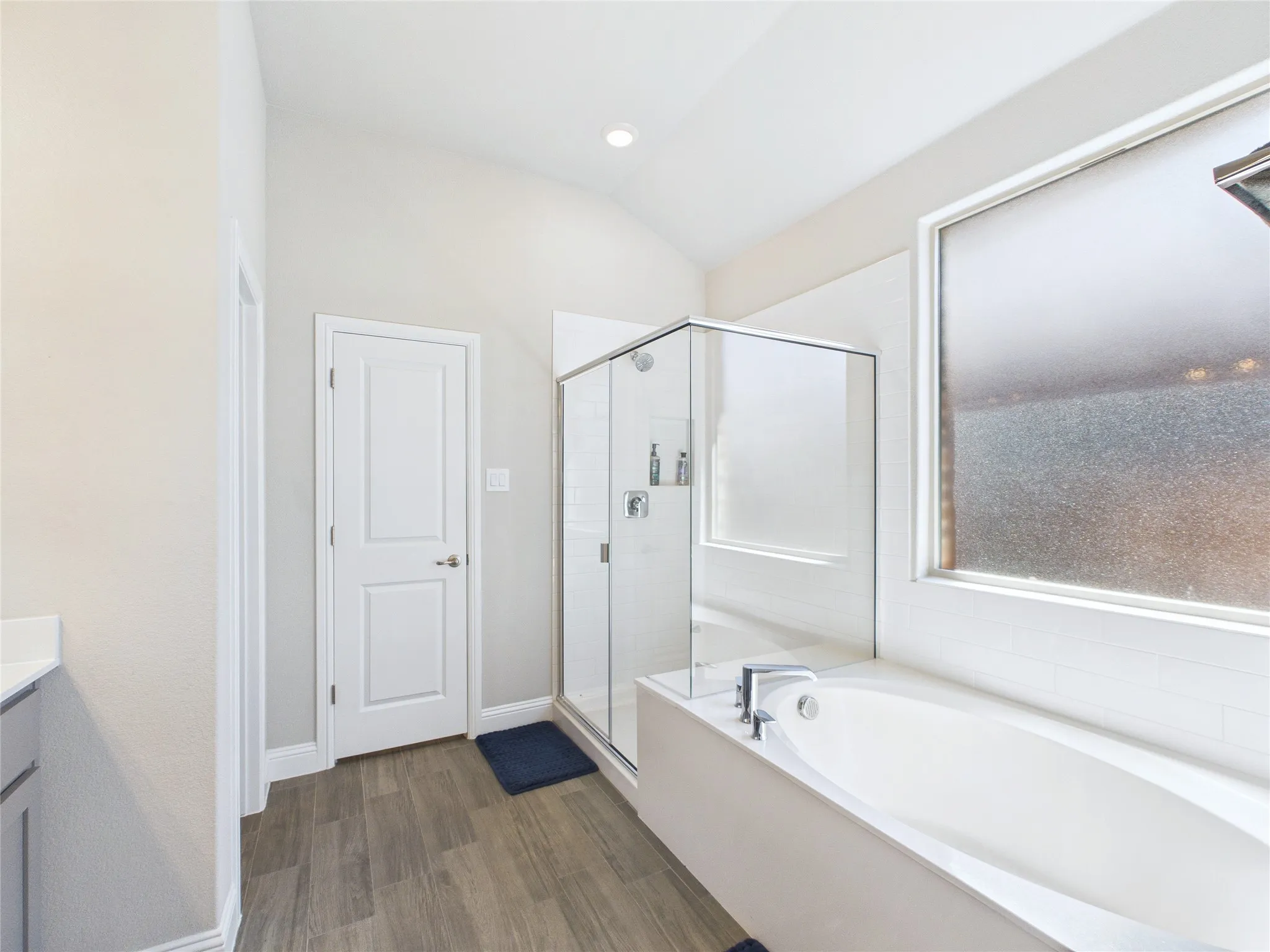 Spacious bathroom featuring a built-in soaking tub, a separate glass-enclosed shower, wood-finish flooring, and a window with privacy glass
