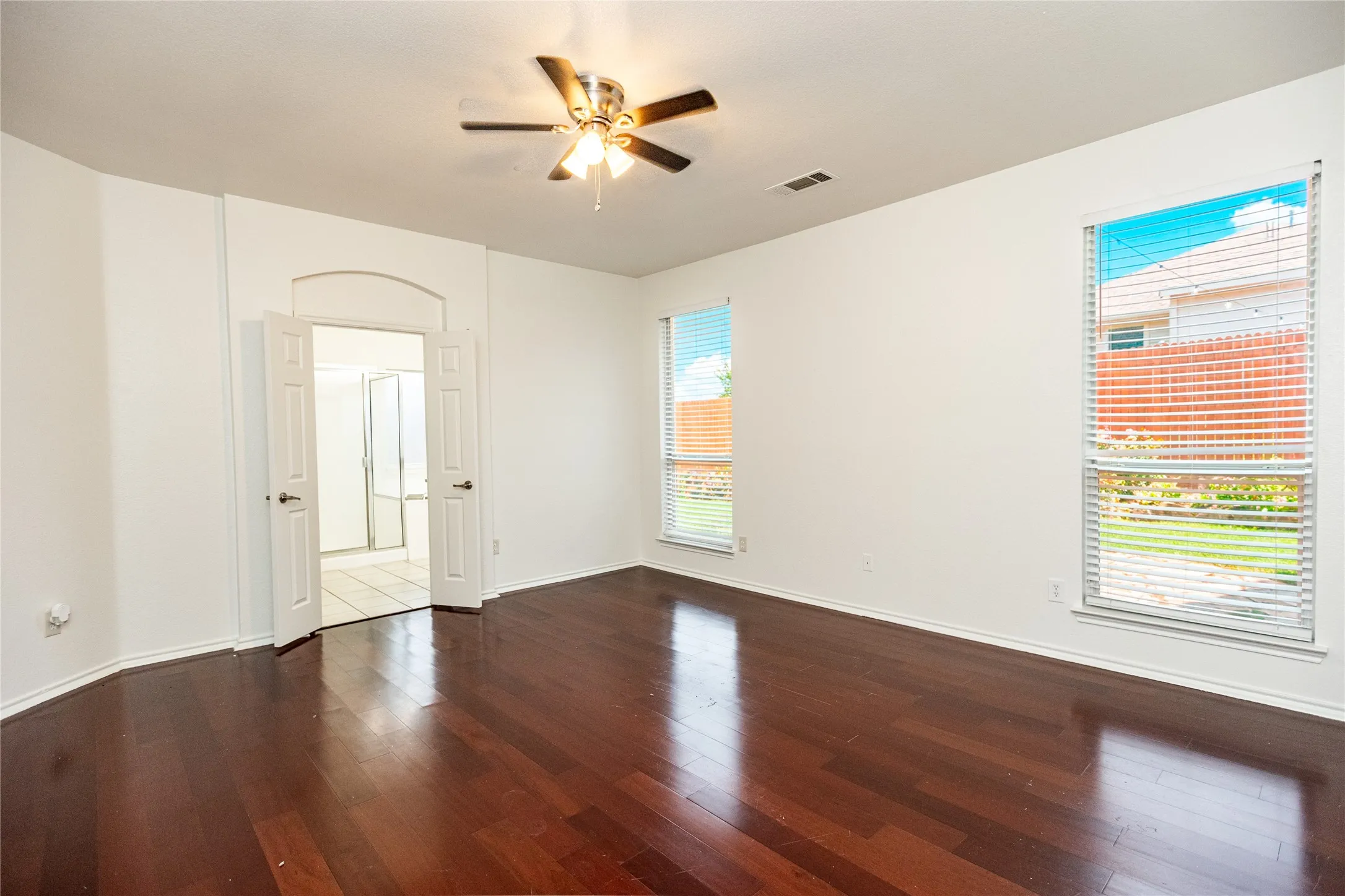 Rich wood-finish flooring, dual windows with blinds, an arched doorway, and an overhead ceiling fan with lighting