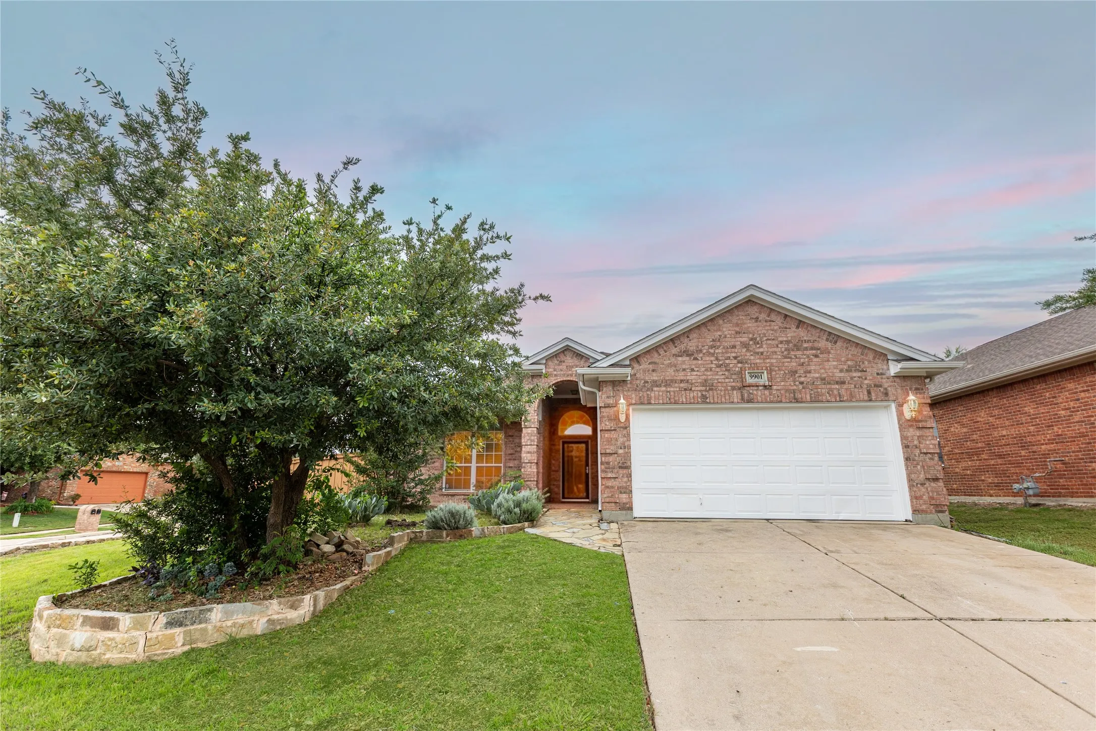 Brick-clad exterior with an arched entry door, two-car garage, concrete driveway, and a stone-lined garden bed