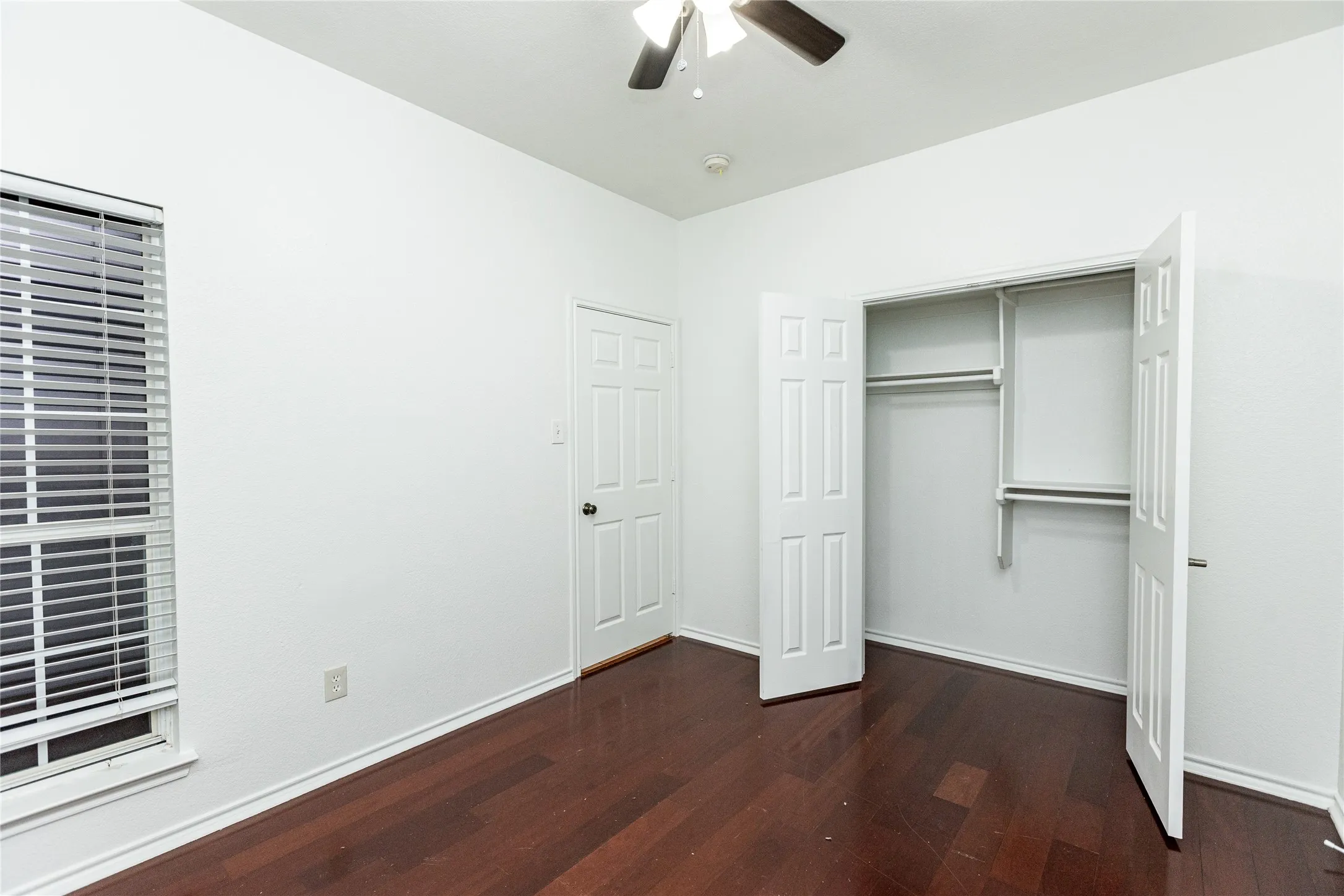 Wood-finish flooring, ceiling fan, window with blinds, paneled door, and a closet with bifold doors and shelving