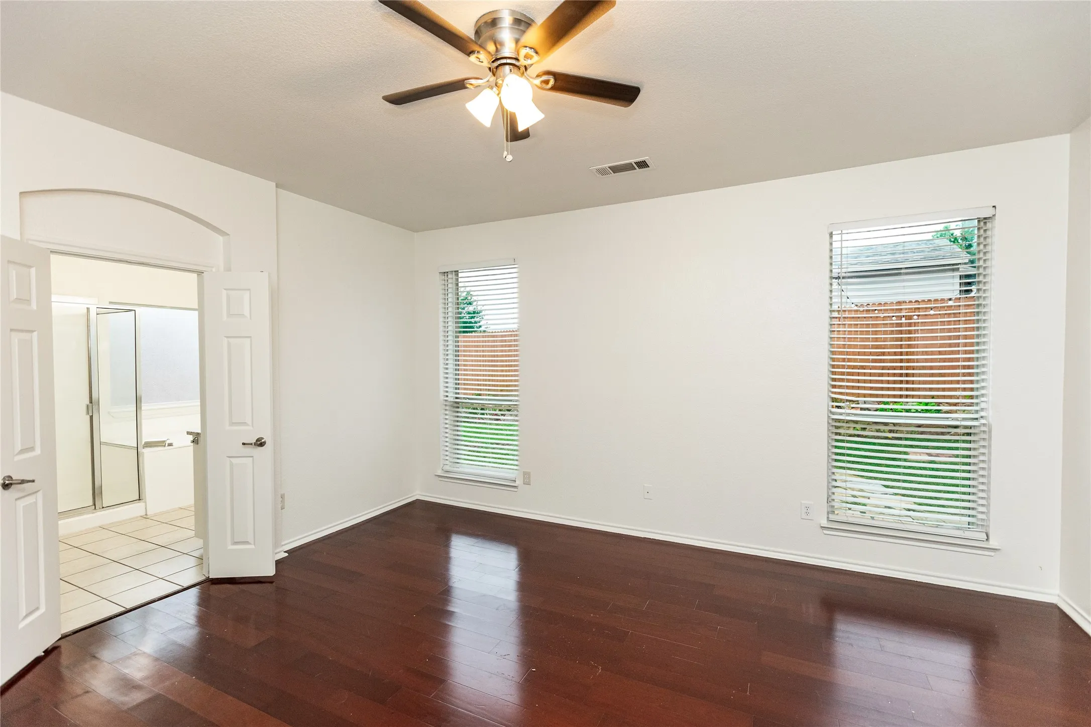 Rich wood-finish flooring, dual windows with blinds, a ceiling fan with integrated lighting, and an arched doorway leading to a bathroom featuring a shower-tub combination