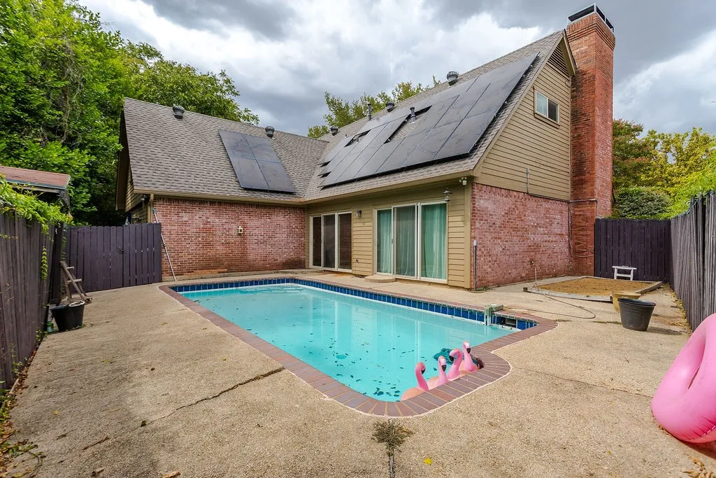 In-ground swimming pool with concrete patio surround, brick and siding exterior, multiple sliding glass doors, brick chimney, and rooftop solar panels