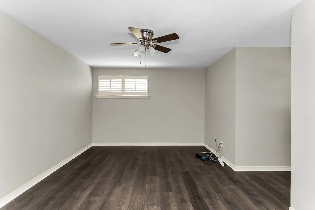 Wood-finish flooring, neutral wall tones, window with plantation shutters, and a ceiling fan with integrated lighting