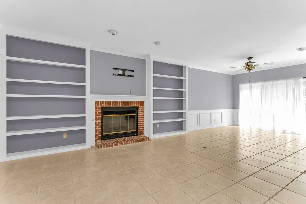 Brick fireplace with a black firebox, flanked by built-in shelving, tile flooring, wainscoting, and a ceiling fan