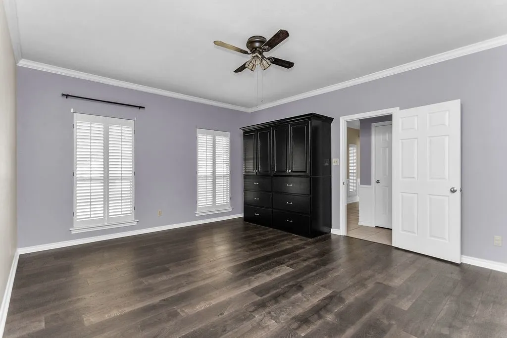 Wood-finish flooring, two windows with plantation shutters, a ceiling fan, crown molding, and a built-in storage unit