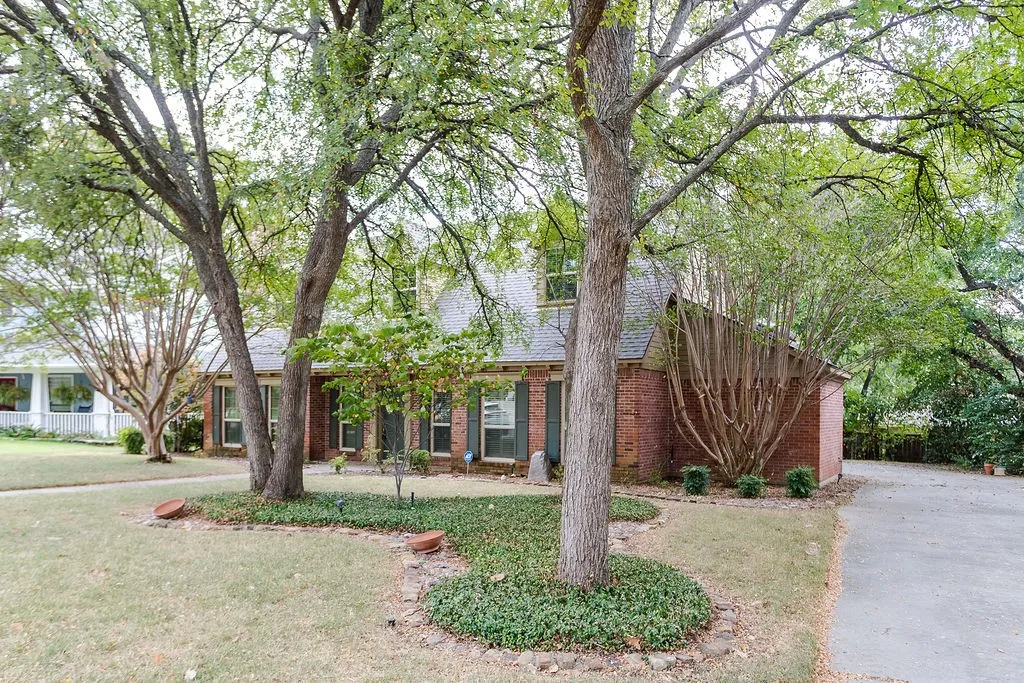 Brick exterior with dormer windows, a covered entry, mature trees, manicured landscaping, and an asphalt driveway