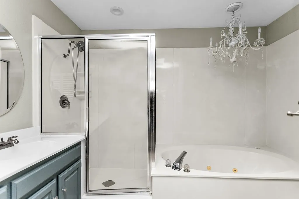 Chandelier lighting, built-in soaking tub, glass-enclosed shower, single vanity with a round mirror, and a gray-painted cabinet