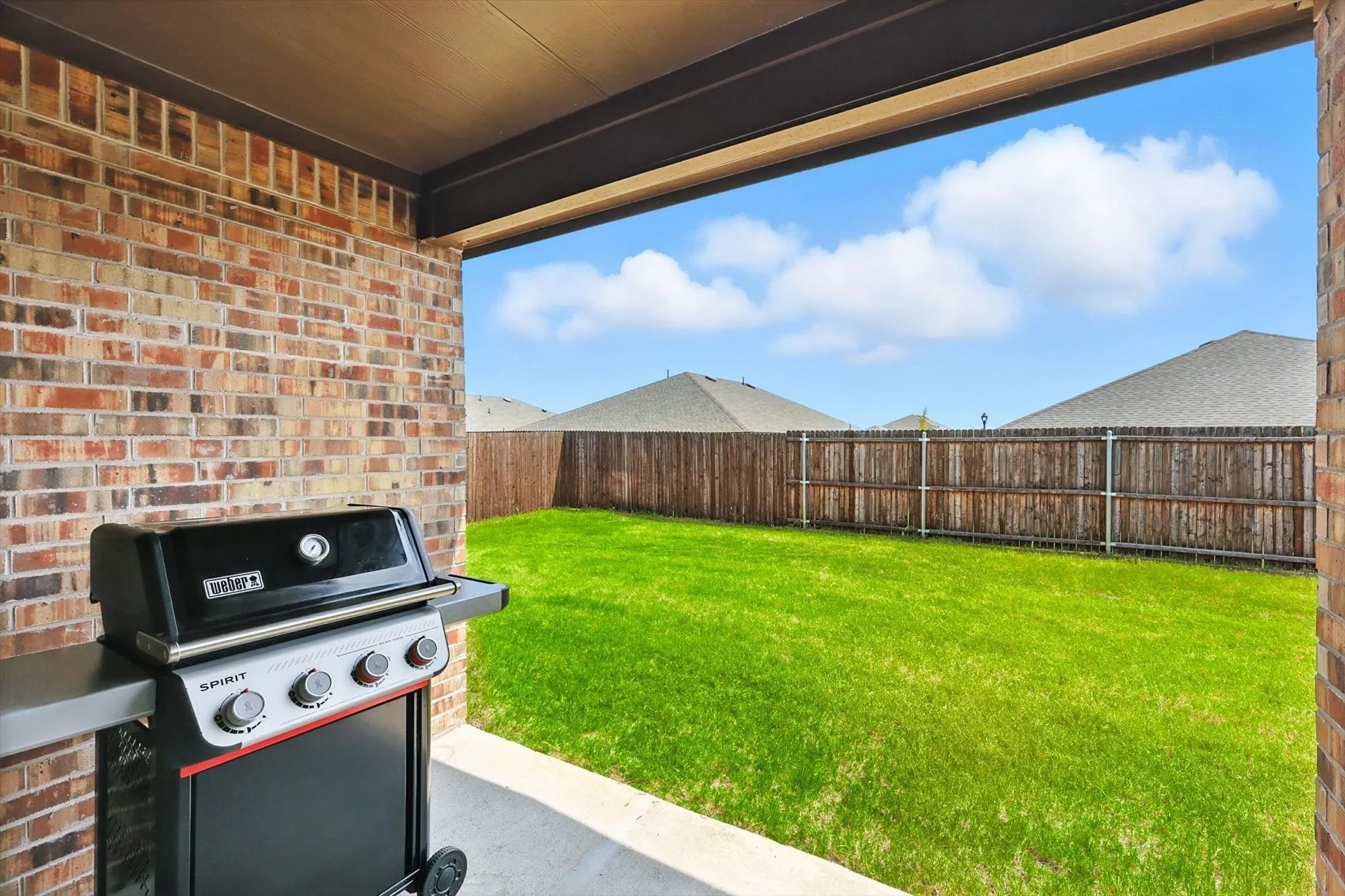 Covered patio with brick wall and concrete flooring, overlooking a green lawn and wood fencing